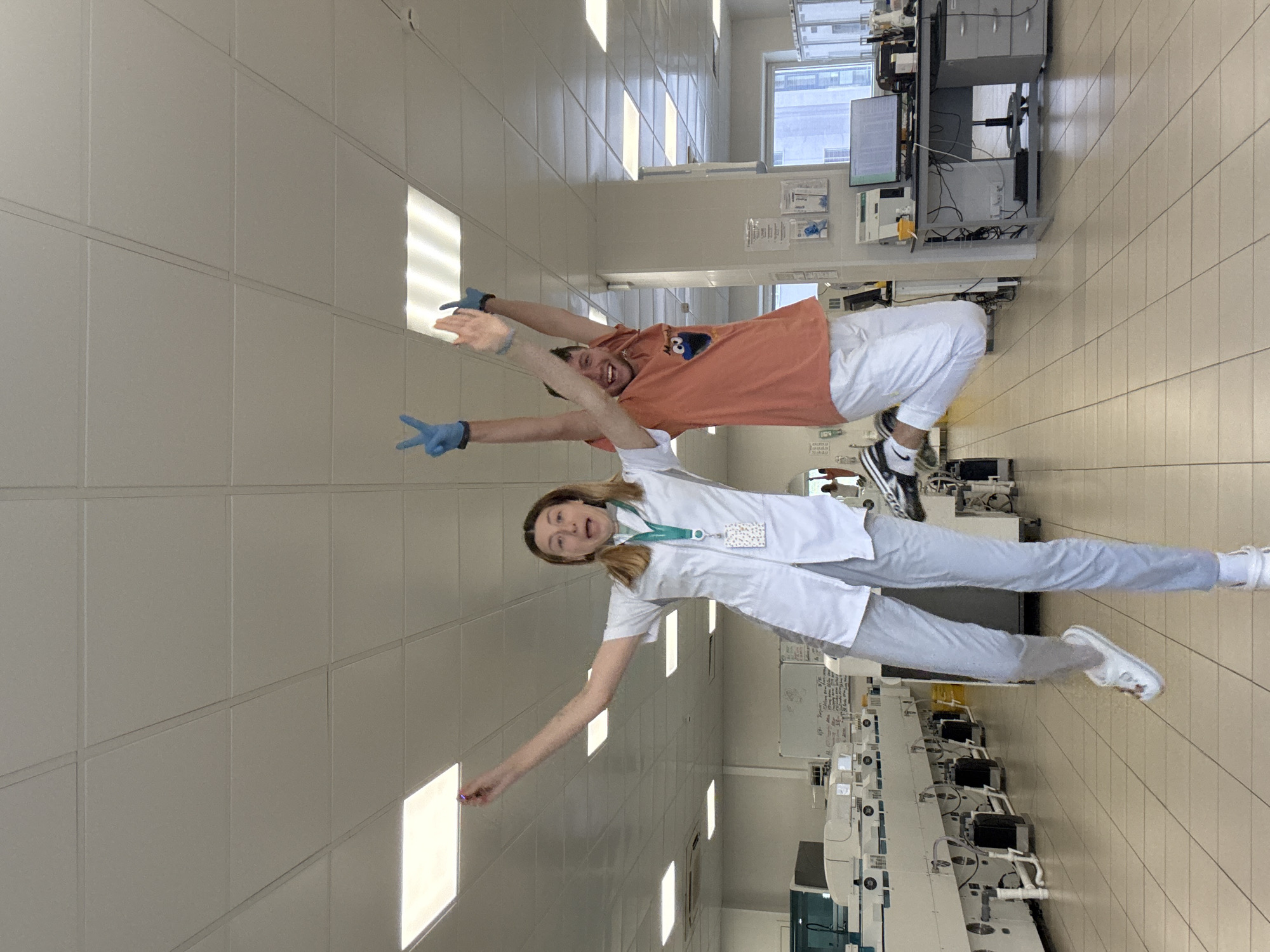 Two individuals, a man and a woman, are posing playfully in a laboratory or cleanroom environment. The man, wearing a light orange t-shirt with a cartoon character and white pants, is in the upper part of the frame. He has blue gloves on both hands, with his right arm extended upwards and his left arm extended outwards, both hands forming a peace sign. He has a wide smile and appears to be looking at the camera. The woman, in the lower part of the frame, wears a white lab coat over grey pants and white sneakers, also sporting blue gloves. Her left arm is outstretched downwards, and her right arm is extended upwards, with her hands also making peace signs. She has an excited expression and seems to be looking towards the camera as well.
The setting is a brightly lit interior with tiled floors and walls, suggestive of a sterile environment like a laboratory or a manufacturing facility. Fluorescent lights are visible on the ceiling. In the background, there are laboratory equipment, computer monitors, and cabinets. The overall atmosphere suggests a moment of lightheartedness and fun amidst a professional setting. The image is captured vertically, with the individuals appearing to be in a jumping or dynamic pose. No specific time of day or weather conditions are discernible. The location context is given as Moscow, Russia, but there are no specific visual cues in the image to confirm this. There is some text visible on a whiteboard in the background, but it is too blurry to be read.