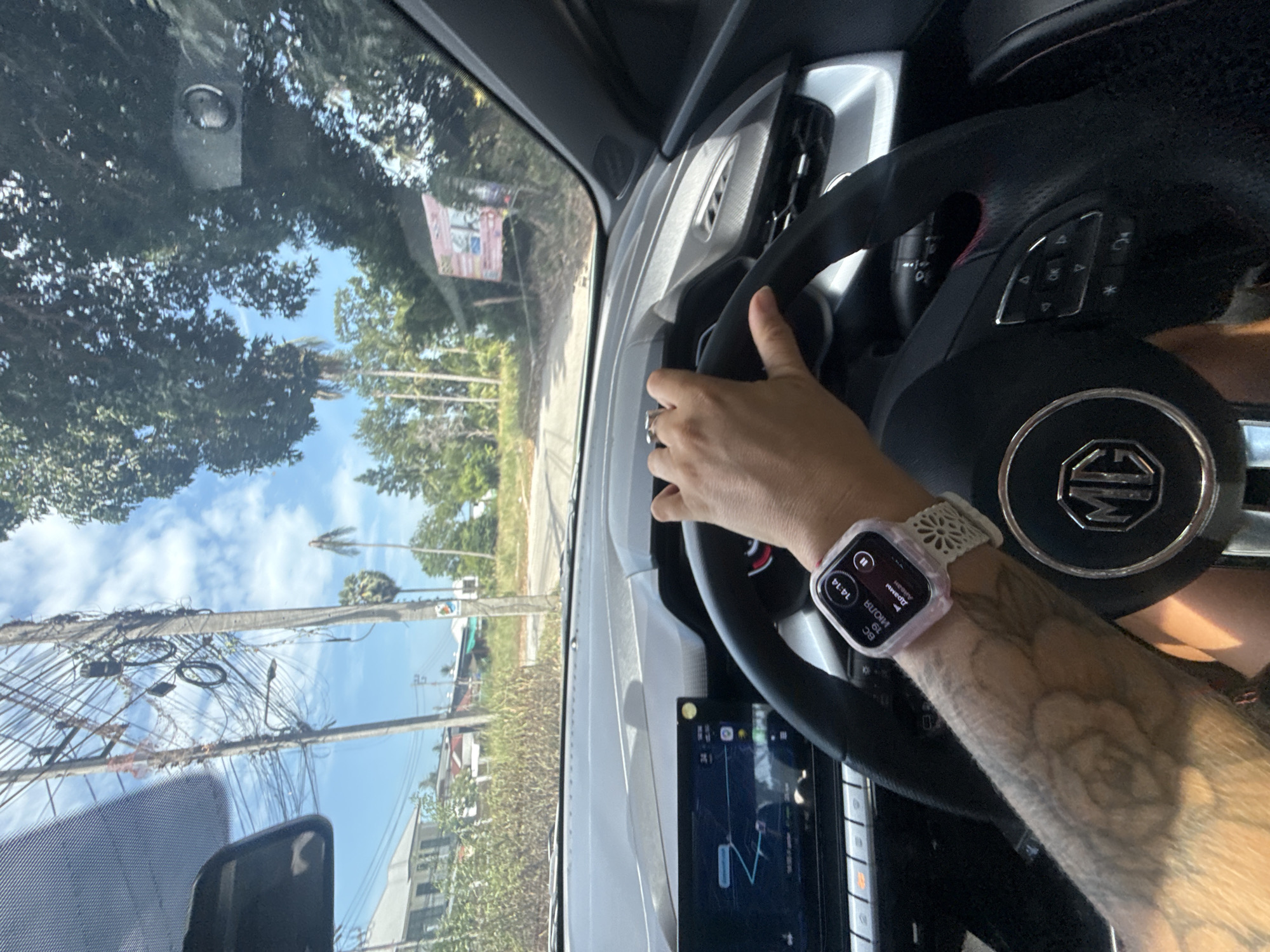 The image shows a view from inside a car, looking out at a road in Ko Samui District, Thailand. A person's hands are visible on the steering wheel. The dashboard displays a navigation screen, and the person is wearing a smartwatch. The exterior view reveals a tropical environment with palm trees, houses, and power lines. The sky is blue with scattered clouds. A sign with Thai text is visible in the distance.