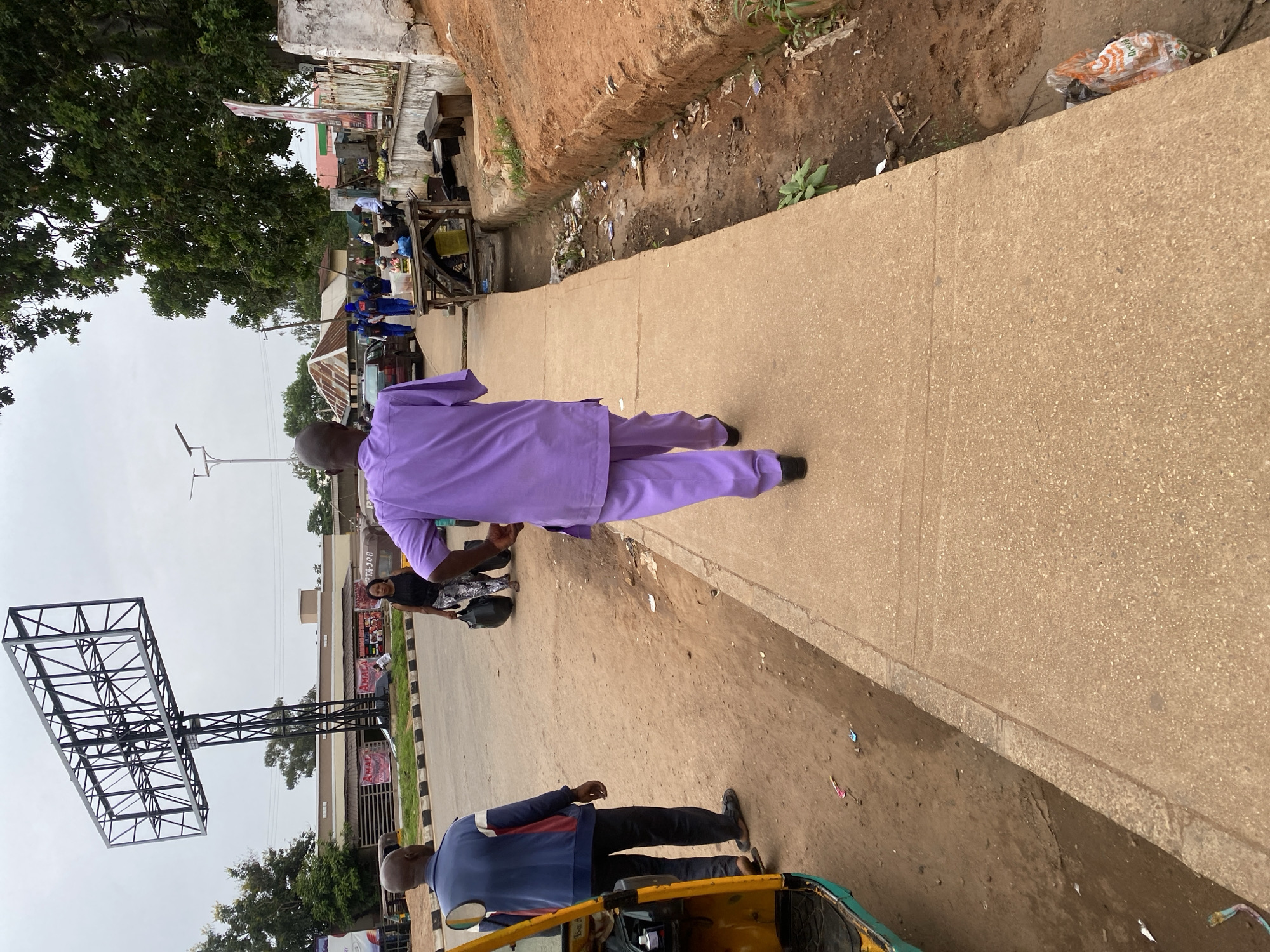 This image depicts a street scene in Jos, Nigeria, likely in a commercial or mixed-use area. In the foreground, a man in a purple traditional outfit is walking away from the camera on a paved sidewalk. To his left, a man in a blue and red jacket is standing on a dirt road, near a yellow auto-rickshaw. Further down the street, a woman carrying two large black bags is walking towards the right side of the frame.
In the background, there is a large, empty billboard structure. Several buildings with signs are visible, including what appears to be a restaurant. A group of people in blue uniforms are walking together on the sidewalk further down. There is a tree-lined area with lush green foliage. The ground is a mix of dirt and pavement, with some scattered litter visible in the drainage ditch alongside the sidewalk. The sky is overcast. The scene suggests a bustling urban environment with pedestrian and vehicular activity.