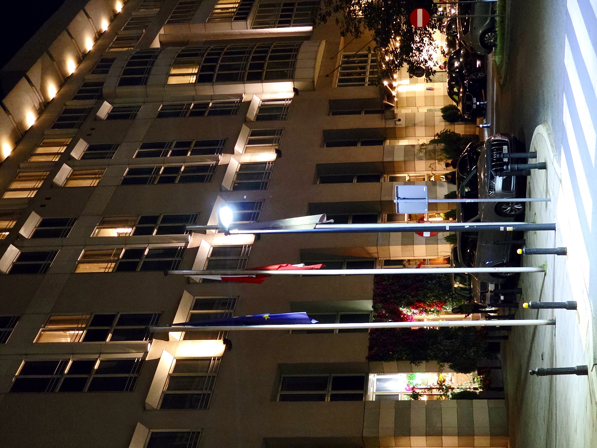 The image captures a nighttime exterior view of a modern hotel building in Warsaw, Poland, likely the Hotel Bristol, based on architectural style and its prominent location. The facade features numerous windows, some illuminated from within, casting warm light onto the exterior. Two flags, one a European Union flag and the other a Polish flag, are prominently displayed on flagpoles in front of the building.
In the foreground, several cars are parked, including a silver Audi SUV with license plate O 888T, a black BMW SUV, and another dark-colored car partially visible. The street in front of the hotel has a pedestrian crossing. Streetlights illuminate the scene, creating a well-lit urban environment. The building's entrance area is decorated with lush greenery and floral arrangements, visible through a large glass window. Several individuals can be seen near the entrance and inside the building. The overall impression is that of a sophisticated and active urban setting at night.