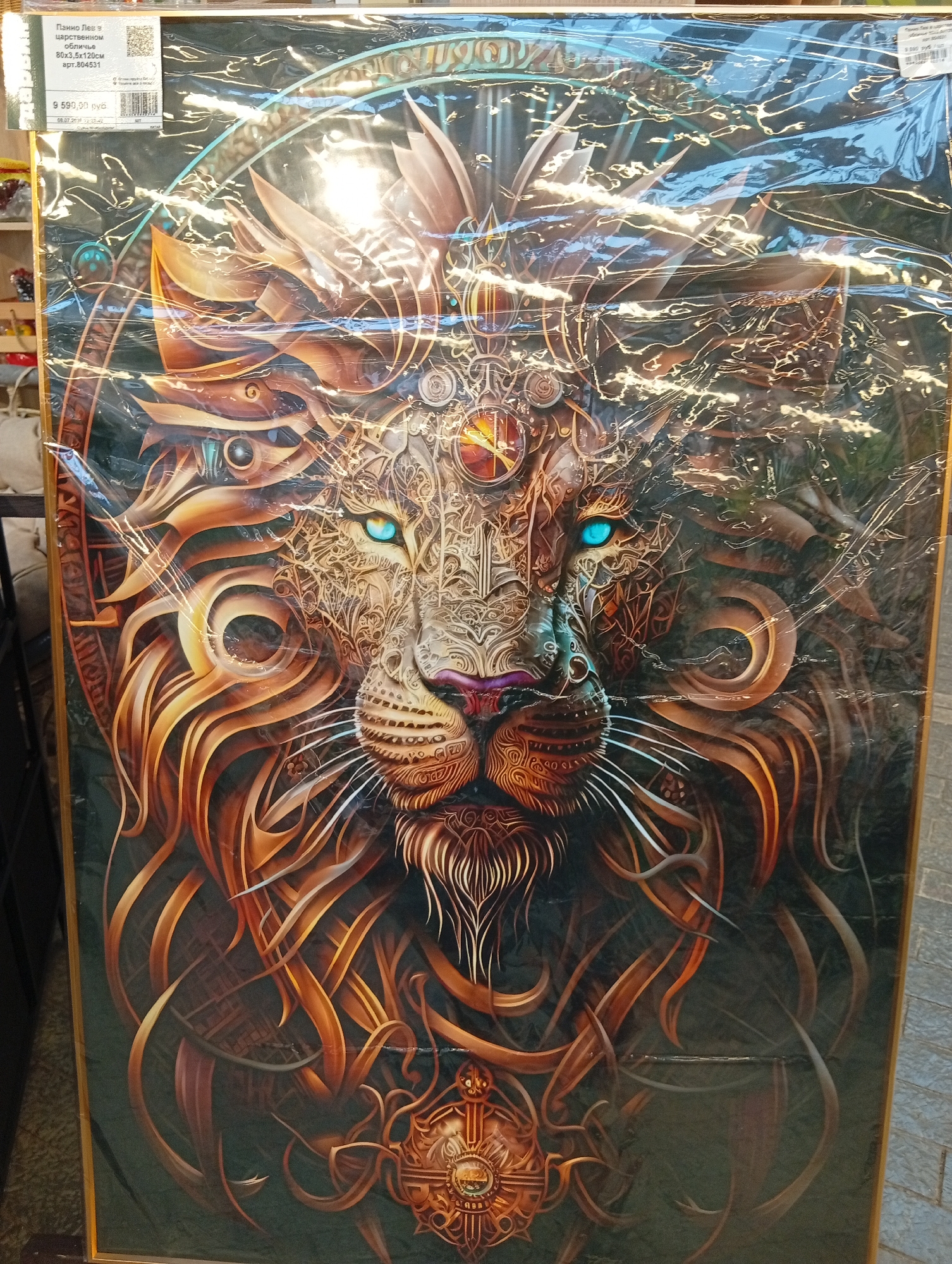 The image shows a framed artwork of a stylized lion's head, predominantly in shades of gold and brown, with striking blue eyes. The lion's mane is intricately detailed with metallic and mechanical elements. The artwork is covered in plastic wrap, and a price tag is visible in the upper left corner. The price tag indicates the item is in Pushkino, Russia, and lists the price as 9,590.00 RUB. The background appears to be a retail environment, possibly a store, with shelves and other items visible. The lighting suggests an indoor setting with overhead illumination, causing some glare on the plastic wrap.