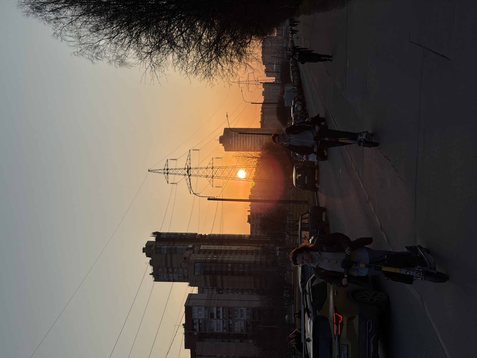 The photograph depicts a street scene in what appears to be Saint Petersburg, Russia. The sun is setting, casting an orange glow across the sky and creating silhouettes of buildings and power lines. On the right side of the image, two individuals are riding electric scooters on a road with parked cars. Other people are walking further down the street. On the left side, apartment buildings are visible. A bare tree is partially visible in the upper left corner of the frame. A car with a license plate is partially visible in the bottom right.