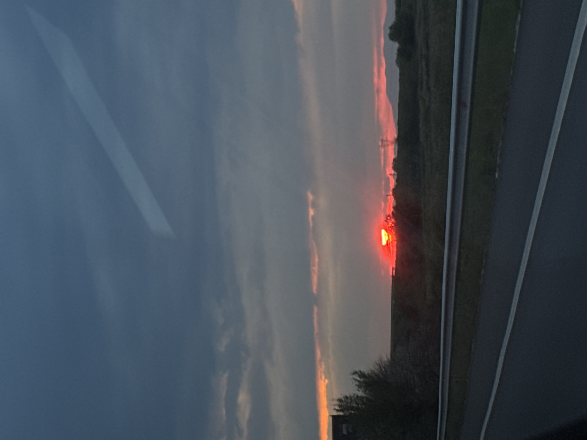 The image, rotated, captures a dramatic sunset scene viewed from inside a moving vehicle. The horizon runs vertically on the right side of the frame in the original orientation, but when viewed correctly, the sun is setting low and vibrant on the right side of the image.
The sky is dominated by a mix of dark grey and lighter clouds, with a striking band of vivid red and orange hues directly above the horizon where the sun is visible. The sun itself appears as an intense, bright red-orange orb, partially obscured by the clouds or atmospheric haze, casting a warm glow across the landscape.
In the foreground, hints of the vehicle's interior are visible on the left side of the image (when correctly oriented), suggesting a window or reflective surface with faint light reflections. The lower part of the scene shows a paved road or highway with clear white lane markings and a silver-colored guardrail or barrier running alongside it. Beyond the road, a dark, flat expanse of rural landscape or field stretches towards the horizon, with sporadic, dark silhouettes of bushes or small trees in the immediate foreground and a distinct power line tower visible in the distance against the glowing sky.
The scene depicts the natural event of sunset, likely experienced during travel along a highway. The time of day is clearly evening, characterized by the intense colors of the setting sun. The weather appears partly cloudy. No people or visible text are present in the image. The location context provided is Malo Konare, Bulgaria, suggesting this rural highway scene is in that region.
