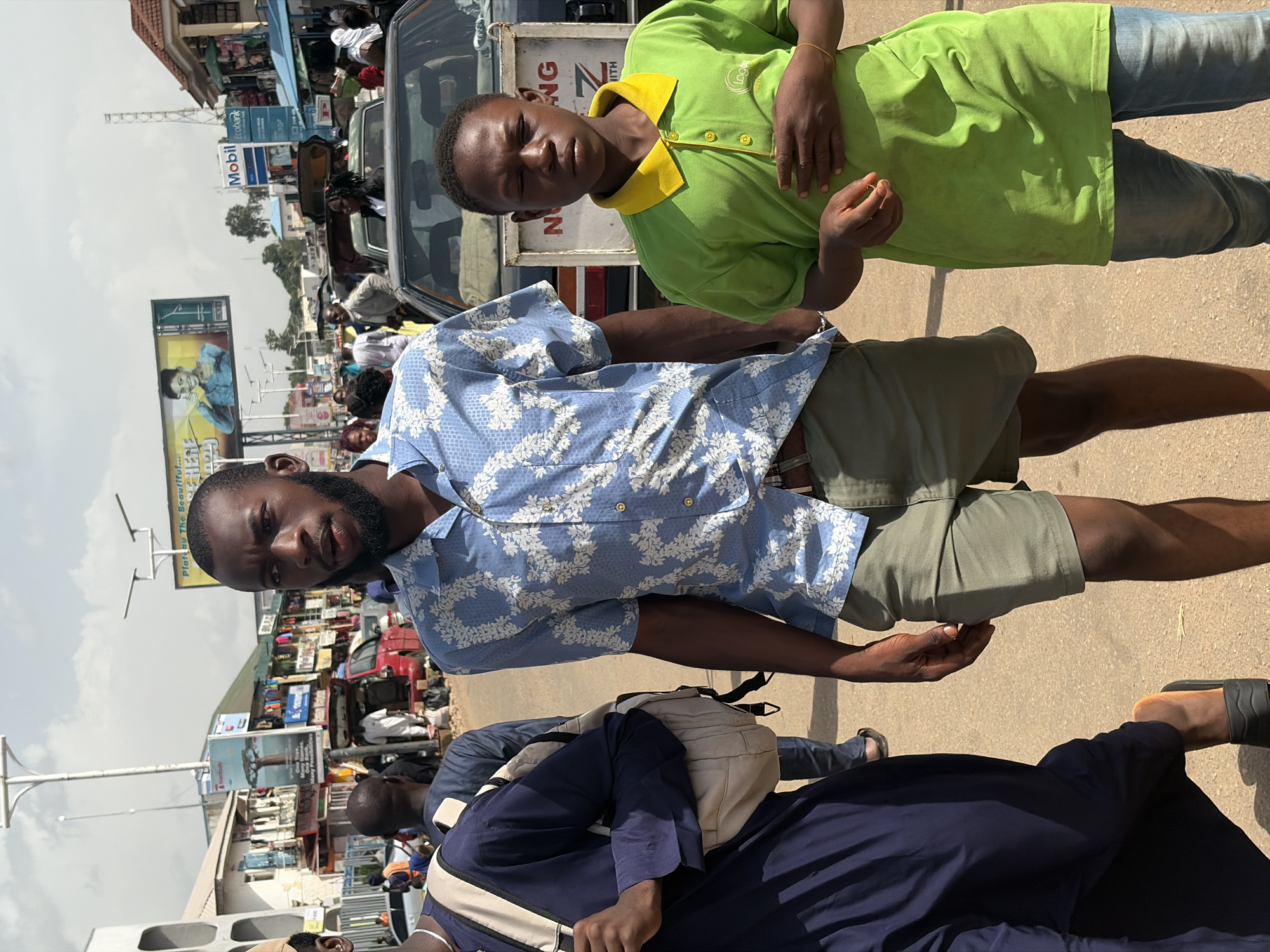 In Jos, Nigeria, a medium shot captures a sunny street scene. The foreground features two young men walking towards the camera. The man on the left wears a navy blue robe and a light-colored backpack. The man on the right is dressed in a light blue short-sleeved shirt with a white floral pattern and olive green shorts. To the right of these two, a younger boy in a bright green polo shirt and blue jeans walks with his hands clasped at his waist. In the background, a busy street unfolds with vehicles, shops, and billboards. A black car is parked on the right side of the frame. One billboard displays an advertisement for Mobil. The sky is bright with scattered clouds.