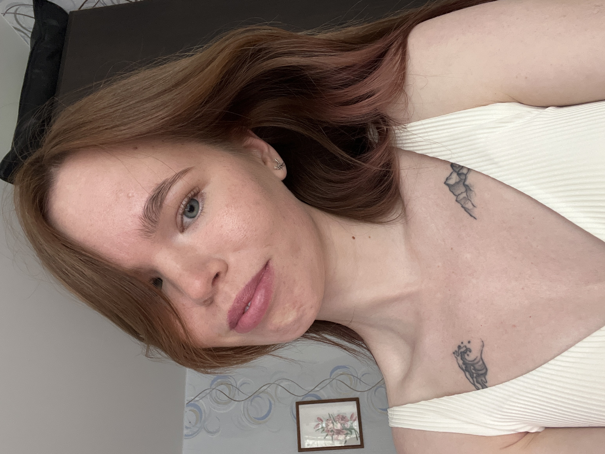 A woman with auburn hair and light skin is facing the camera. She has blue eyes and wears pink lipstick. Her hair has pink highlights. She wears a white, ribbed top. Two tattoos are visible on her upper chest and shoulder area. The background includes a bed, a patterned wall, and a framed picture of flowers. While I can describe the scene, I can not confirm the location in the city of Chaikovsky, Russia.
