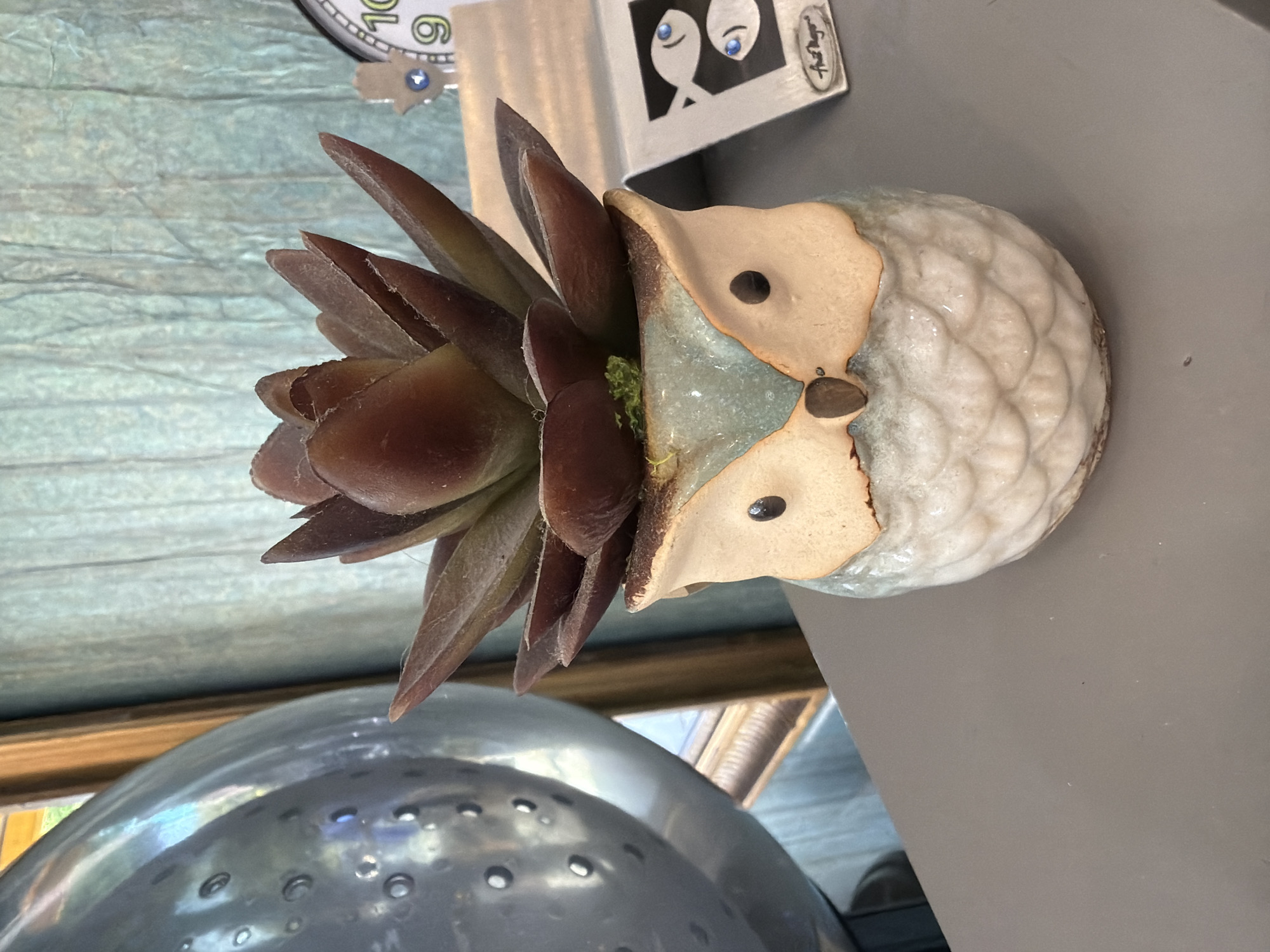 An artificial succulent plant sits in a ceramic owl-shaped planter. The owl planter has a textured, off-white body with a light blue-green glaze around its face. It features tan "feather" markings and black eyes. The succulent has thick, pointed, dark reddish-brown leaves. The planter is positioned on a flat, gray surface. In the background, a textured, teal-colored fabric hangs vertically. To the left of the planter, a large, reflective, spherical object with circular indentations is partially visible. To the right, a small, silver stand holds a rectangular card with an image of two cartoonish fish and a signature, and behind that, a small clock face with Arabic numerals and what appears to be a hamsa hand charm is visible. The overall scene is an indoor setting, likely a home or retail environment.