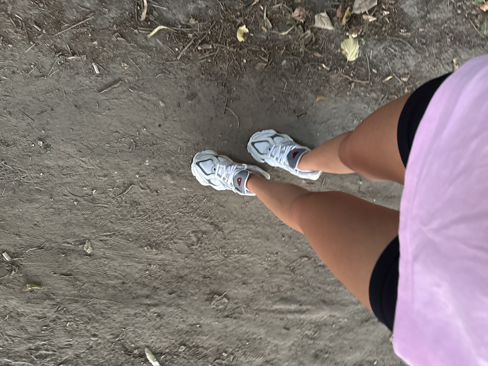 A person wearing white athletic shoes, black shorts, and a pink shirt is walking on a dirt path. The path is covered with loose dirt, small stones, and dried leaves. The image is taken from a high-angle perspective, looking down at the person's legs and feet. The lighting suggests it is daytime. The specific location in Chișinău, Moldova, is not discernible from the visual information present in the image.
