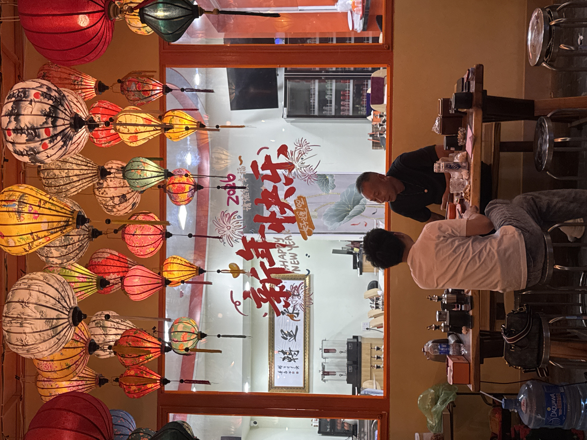 The image depicts an indoor scene in what appears to be a commercial establishment. Two individuals are visible: one, dressed in a black shirt, is standing while the other, wearing a white shirt and grey pants, is sitting. They appear to be engaged in conversation or possibly interacting with items on a wooden counter.
Behind them, a window showcases a decorative display including Chinese characters, specifically the phrase "Happy New Year," and other ornate designs. Through the window reflection, a television and a refrigerated display case filled with beverages are discernible.
Hanging from the ceiling are multiple traditional Vietnamese lanterns of varying colors and patterns. To the right of the seated individual, stacks of glasses and containers, and bottled water, can be seen along with what seems to be a black handbag. The overall environment suggests an establishment located in Đà Nẵng, Vietnam.