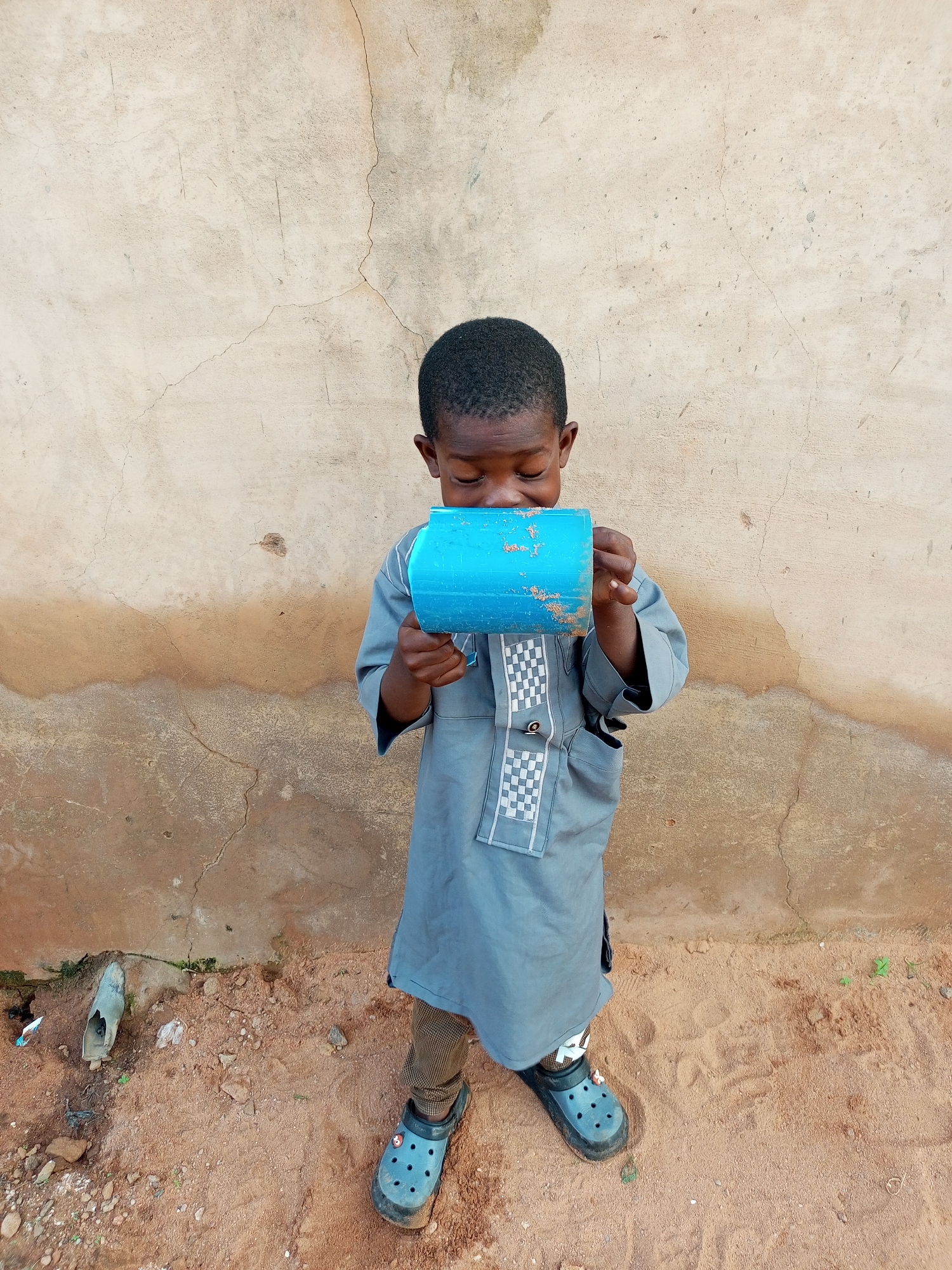 A young boy of African descent, dressed in a grey traditional tunic, stands in front of a textured, light-brown wall in Tempure, Nigeria. He is holding a blue cylindrical object to his mouth, appearing to drink from it. His eyes are closed, and his head is tilted slightly downwards. The ground beneath him is sandy and scattered with small stones and debris. One foot is clad in a black clog, while the other is partially obscured, also appearing to be a black clog. The wall exhibits cracks and has a darker, damp-looking stain running horizontally across its lower half.