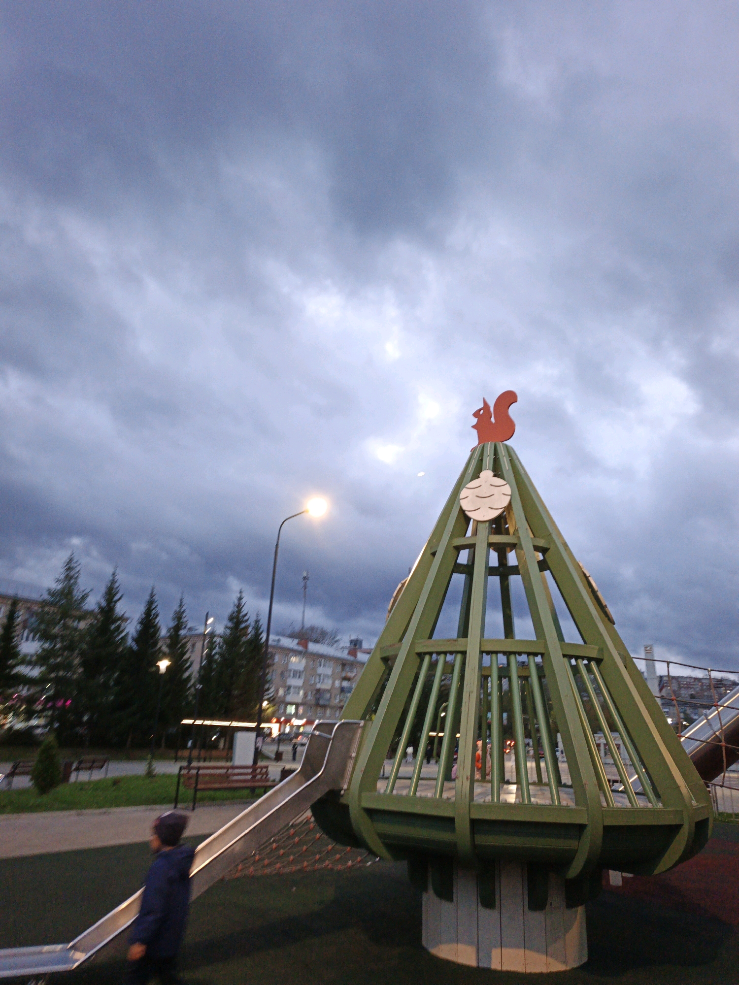 The image depicts a playground in Zlatoust, Russia, under a cloudy sky. A distinctive green, cone-shaped play structure, topped with a red squirrel figurine and decorative circles, dominates the foreground. To the left, a child in a dark blue hooded jacket stands on a metal slide. In the background, evergreen trees, buildings, and a street lamp are visible. The lighting suggests it is late afternoon or early evening, with the sky a muted gray.