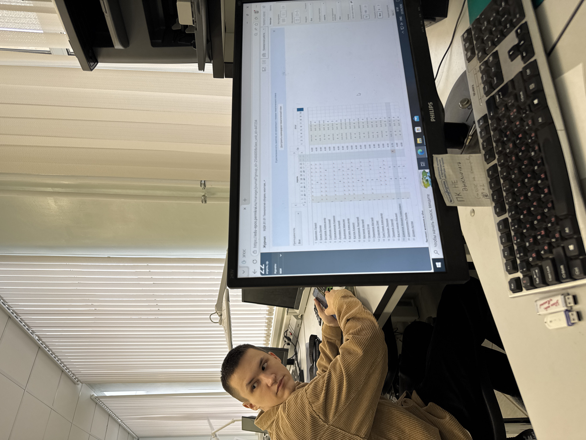 A young male, possibly a student or educator, is seated at a desk in what appears to be an office or classroom in Perm, Russia. He is wearing a light brown corduroy jacket and holding a smartphone, looking directly at the camera with a neutral expression.
The primary focus is a Philips computer monitor displaying a Russian educational portal ("edu-epos.permkrai.ru"). The screen shows a digital journal or gradebook for "МДК.01.01 Технология сборки, монтаж" (Assembly, installation Technology), listing names like "Бабенко Алина" and "Зырянов Максим" with associated numeric data, likely grades or progress. Visible text on the screen also includes "Экспорт журнала в xml" (Export journal to xml), "Достроить календарно-тематический план" (Complete the calendar-thematic plan), and "В расписании есть занятия" (There are classes in the schedule). The top right corner of the monitor displays "8:50 ENG 22.04.2020," indicating the time and date.
To the right of the monitor, a black Sven keyboard and a partially visible mouse are on the desk. A handwritten note is taped to the monitor stand with the message "ПК НЕ Выключать" (DO NOT Turn Off PC) and a smaller note below that seems to be an internal reference, "для БОТС на Внедр-гору." A small sticker also mentions "профилактика анаферонастуды и гриппа" (prevention of anaferon cold and flu). Two USB drives are plugged into a device on the desk next to the keyboard. In the background, white or beige horizontal window blinds are visible, suggesting an indoor setting with natural light. The overall scene suggests a routine workday or study session.