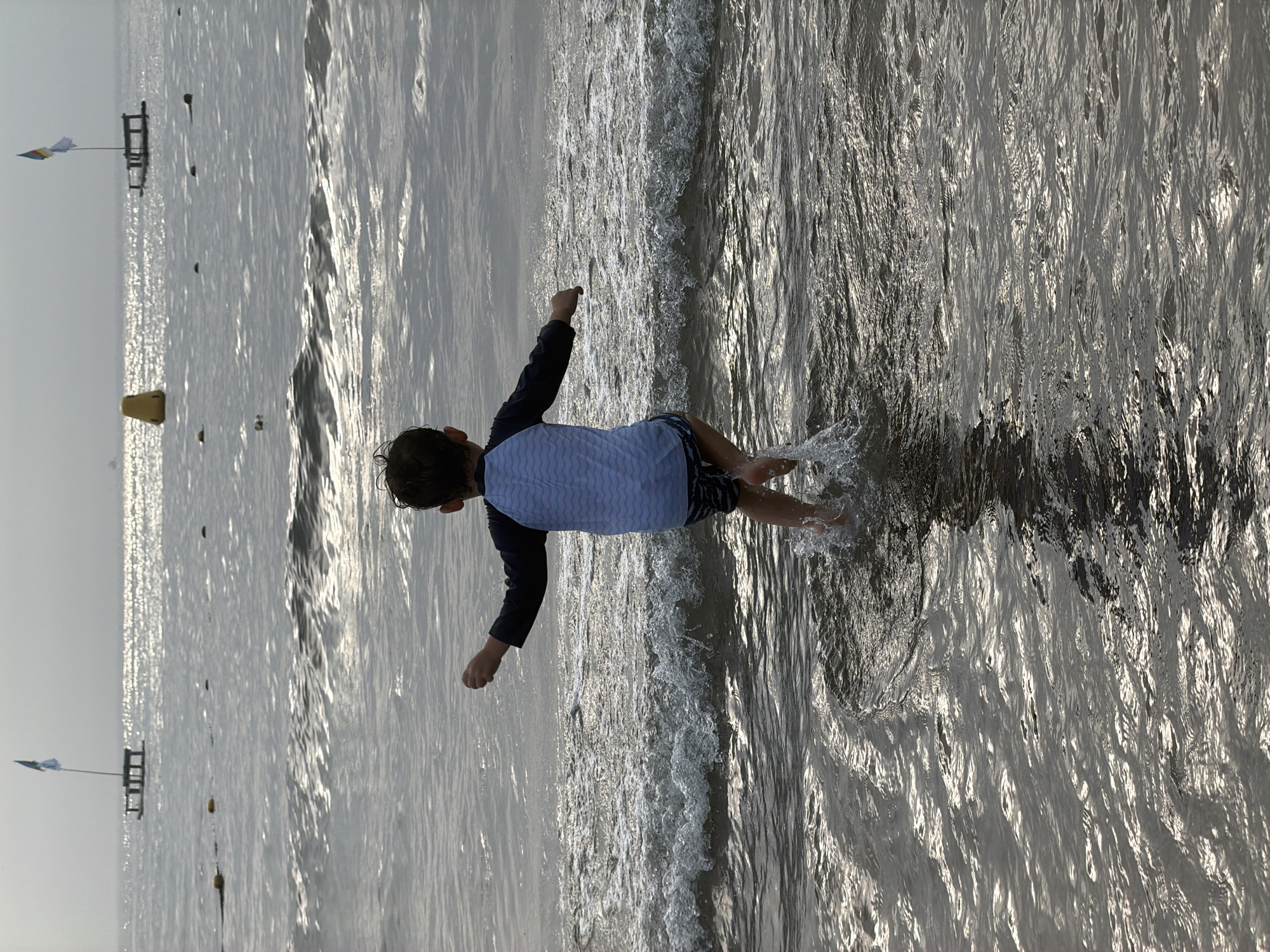 A young child, seen from behind, runs into the ocean at a beach, splashing water. The child wears a long-sleeved rash guard with a light blue wave pattern on the front and dark shorts. The ocean is choppy with moderate waves breaking. In the distance, two floating platforms with flags are visible, along with buoys marking a swimming area. The sky is overcast and the sunlight reflects off the water. This scene appears to be at Hyeopjae Beach, Jeju City, South Korea, known for its shallow, calm waters and white sand.