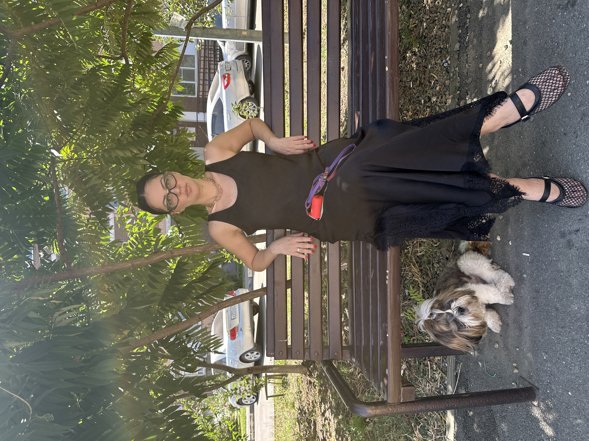 A full shot captures a woman and a dog on a park bench under the shade of trees. The woman, with dark hair pulled back and wearing glasses, is seated on the right side of the bench, dressed in a long black sleeveless dress with lace trim and mesh flats. Her left arm rests on the back of the bench, and a red and purple dog leash lies across her lap. To her left, a small, fluffy Shih Tzu dog sits on the ground beneath the bench. The bench itself is made of dark brown wooden slats and a metal frame. In the background, silver cars are parked on a street adjacent to the park, and a multi-story building is visible. The scene is lit by natural sunlight, creating dappled shadows from the trees.