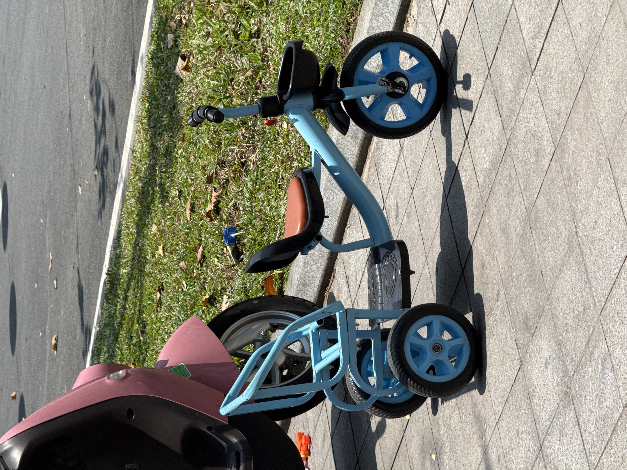 The image captures a light blue children's tricycle parked on a gray paved sidewalk, with a portion of a pink scooter or motorbike visible on the far left. The tricycle, which has black handlebars and a brown seat, features three wheels with blue spokes and a black plastic platform with a footprint design between its rear wheels.
Adjacent to the sidewalk is a narrow strip of green grass dotted with dry leaves, separating the paved area from an asphalt road. The road has a clear white line marking its edge. Strong, defined shadows from the tricycle and other objects fall across the sidewalk and grass, indicating bright, direct sunlight, suggesting it is midday or early afternoon. The weather appears clear and sunny.
The pink scooter, positioned partially in the foreground on the left, shows a light-colored headlamp and a small green sticker on its body, though the text on the sticker is unreadable. No people are present, and the vehicles appear to be stationary, suggesting a paused or unattended scene in an outdoor setting in Phú Quốc, Vietnam.