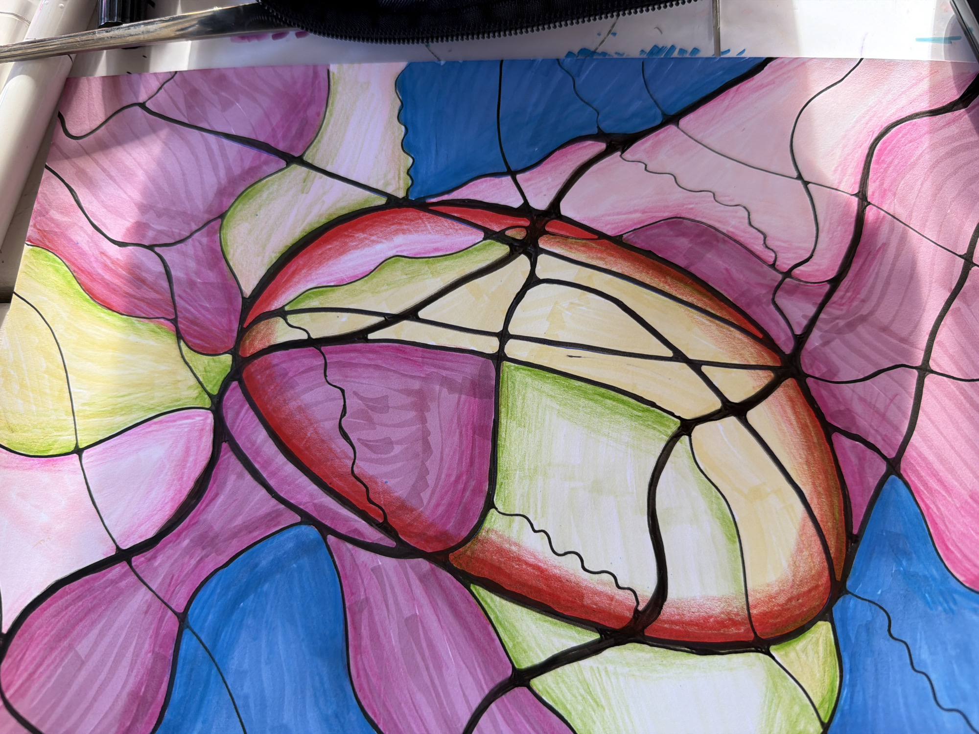 The image shows a colorful abstract drawing on white paper. The drawing consists of organic shapes outlined in black and filled with pink, purple, blue, yellow, green, and red hues. The shapes are arranged in a somewhat radial pattern, with a central, elongated oval shape. The overall impression is of a vibrant and flowing design. The scene is indoors and the drawing is illuminated by natural light.
There is no information in the image to indicate that the photo was taken in Budva, Montenegro.