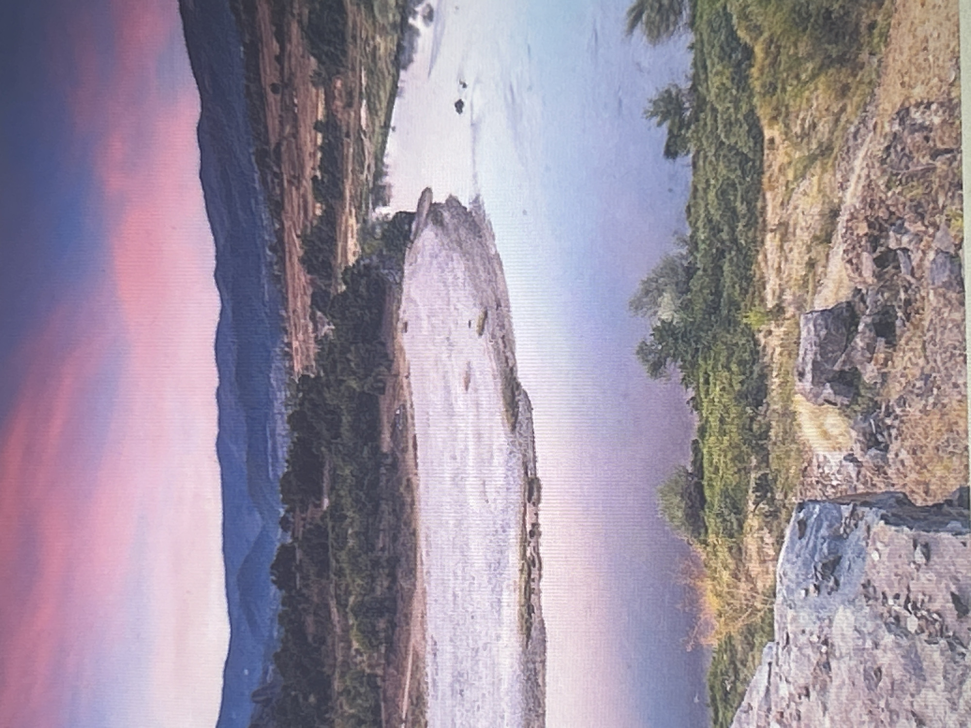 The image shows a picturesque landscape with a river flowing through a valley. In the foreground, there are large rocks and dry vegetation. The river curves through a sandy bank, bordered by trees and green foliage. The middle ground features more trees and rolling hills. In the background, a mountain range is visible against a sky filled with soft pink and purple clouds, suggesting a sunset or sunrise. The overall scene is serene and natural, with no people or man-made structures clearly identifiable. The location is not specified in the city of Sofia, Bulgaria.