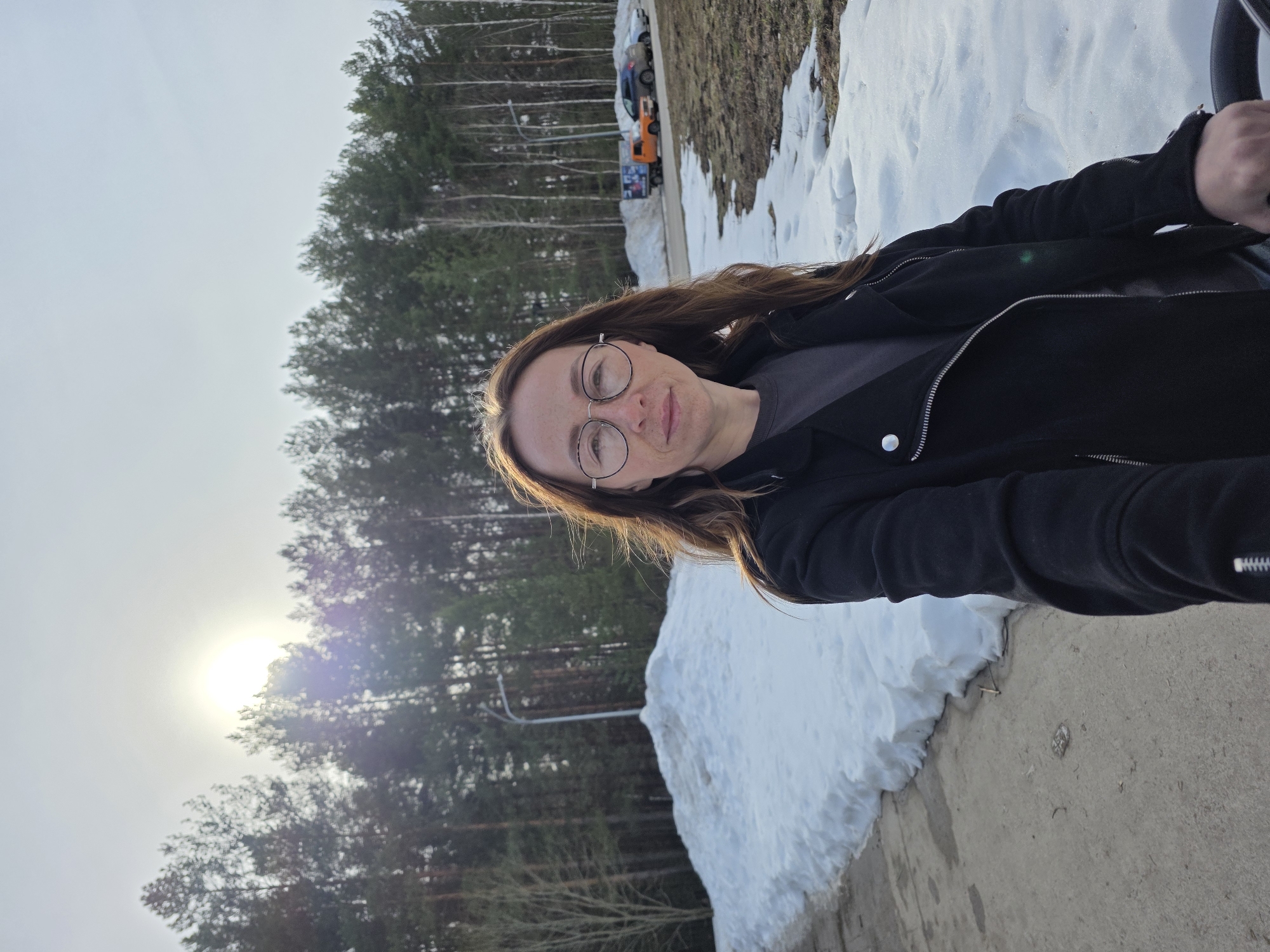 A person is standing on a concrete surface with snow piles beside them. Behind the person is a forest with tall trees, and in the distance, there are parked vehicles. The sun is visible through the trees, creating a hazy sky.
While the exact location within Chaikovsky, Russia cannot be determined with certainty from the image alone, the presence of the forest, snow, and urban elements suggests a location in a park or green space near the edge of the city.