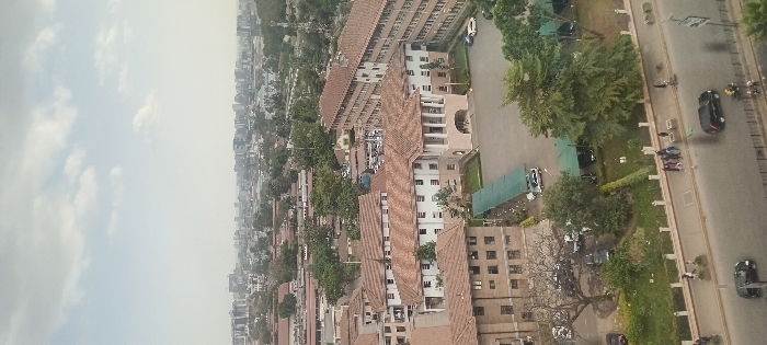 An aerial view depicts a cityscape under a cloudy sky. Buildings with red-tiled roofs stand prominently amidst lush greenery. A road runs along the right side, populated with cars and motorcycles. Further in the distance, the city extends with more buildings, gradually fading into the horizon. Trees are scattered throughout the scene, adding a natural element to the urban landscape.