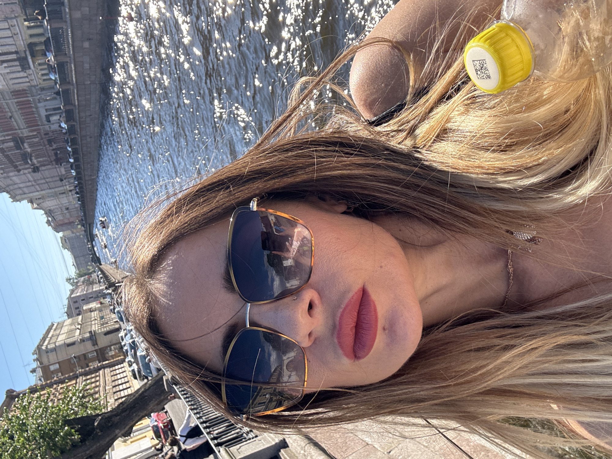 This media file shows a young woman taking a selfie outdoors, possibly on a boat or by a canal, given the reflections of buildings in the water and the presence of a body of water. She is wearing sunglasses and has makeup on her lips. Her hair is long and blonde, and she is holding a clear plastic bottle with a yellow cap. The scene appears to be set during the daytime, under bright sunlight, as indicated by the glare on the water and the shadows. The background shows a line of buildings with architectural details, suggesting an urban environment. There is a QR code visible on the yellow cap of the bottle. The overall mood seems relaxed and casual, typical of a tourist or someone enjoying a leisurely day out. The location is identified as Saint Petersburg, Russia, which aligns with the canal-side setting and the architectural style of the buildings.