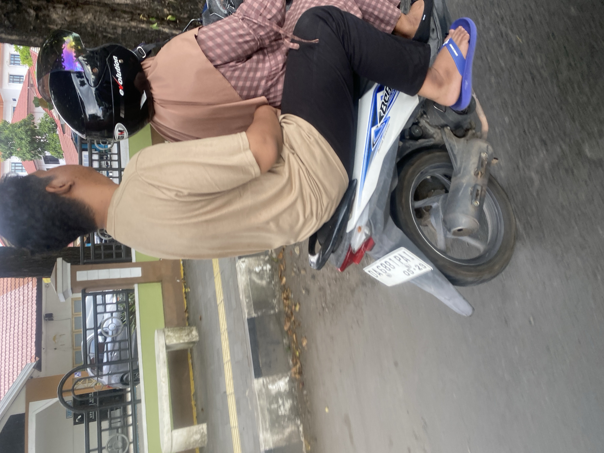 Two individuals are riding on a motorcycle in Banjarbaru, Indonesia. The person in the foreground, who is facing away from the camera, is wearing a tan t-shirt and black pants. The person behind them is wearing a brown hijab and a plaid dress, and has a black helmet with a reflective visor. The motorcycle is white with blue and black accents. The license plate of the motorcycle is visible and reads "DA6881PA1 05-29". They are on a paved road with a sidewalk to their left. In the background, there is a building with a red tiled roof and a tree.