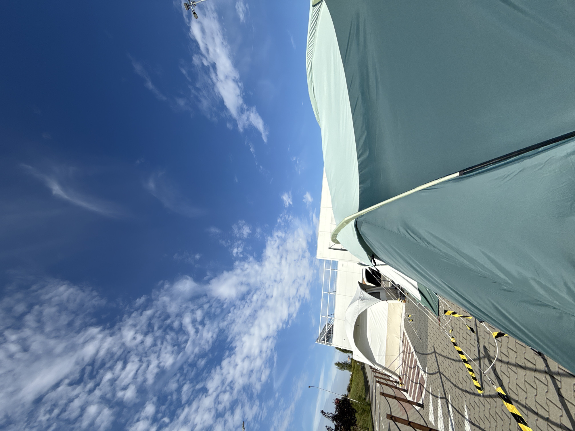 The image depicts an outdoor scene on a bright, cloudy day, likely in the vicinity of Bielany Wrocławskie, Poland. In the foreground, a large, teal-colored tent dominates the right side of the frame, its fabric stretched taut and curving upwards. The tent appears to be set up on a paved surface composed of interlocking concrete blocks.
Behind the teal tent, a white, domed tent structure is visible, with an arched entrance. This structure is adjacent to a modern, white building with a visible white railing along its upper level, suggesting it might be a temporary setup or an outdoor event space.
A pathway or queueing area is marked by wooden posts and rope barriers on the left side of the image, with yellow and black striped caution tape strung between poles. The ground is further delineated by patterned shadows cast by the tents and barriers. In the background, there is a grassy slope with some trees, and a street lamp or light pole with a camera mounted on top is visible against the blue sky dotted with white, wispy clouds. The overall impression is of a functional outdoor setup, possibly for an event, testing facility, or temporary accommodation, under a clear sky.