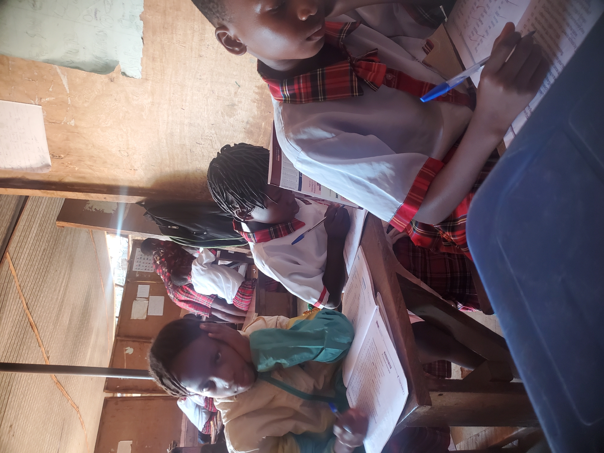 Several students are seated at wooden desks in a classroom in Ukwu-Abwa, Nigeria. The students are wearing school uniforms that consist of white shirts with red and black plaid accents. They appear to be working on assignments, writing on papers with blue pens. The room is enclosed by wooden walls. Some papers are pinned to the walls. Natural light enters the room through an opening in the wall.