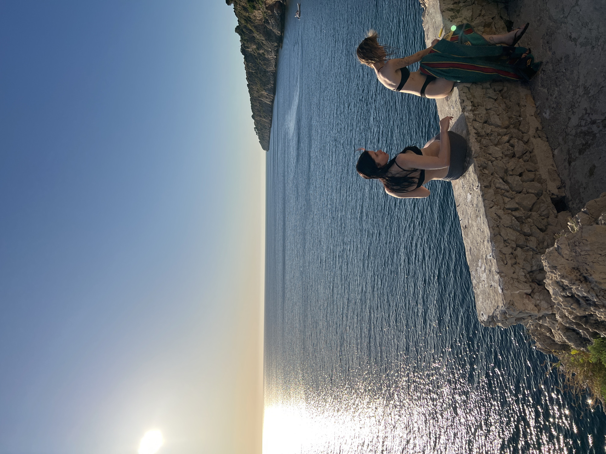 Two women are sitting on the edge of a rocky cliff overlooking the sea. The woman on the left has dark hair and is wearing a black bikini top and grey shorts. She is looking down at the water. The woman on the right has blonde hair and is wearing a black bikini top. She has a colorful patterned sarong draped over her legs. She is looking out towards the sea.
The scene appears to be set during the late afternoon or early evening, as indicated by the low angle of the sun and the warm glow on the water. The sun is reflecting brightly off the surface of the ocean, creating a shimmering effect. The water is a deep blue, and there is a rocky coastline with vegetation on the left side of the image. In the distance, a small boat can be seen on the water. The overall mood of the image is serene and peaceful, with a sense of adventure and freedom. The location appears to be coastal, likely in a Mediterranean region given the context of Terrasini, Italy. There is no visible text in the image.