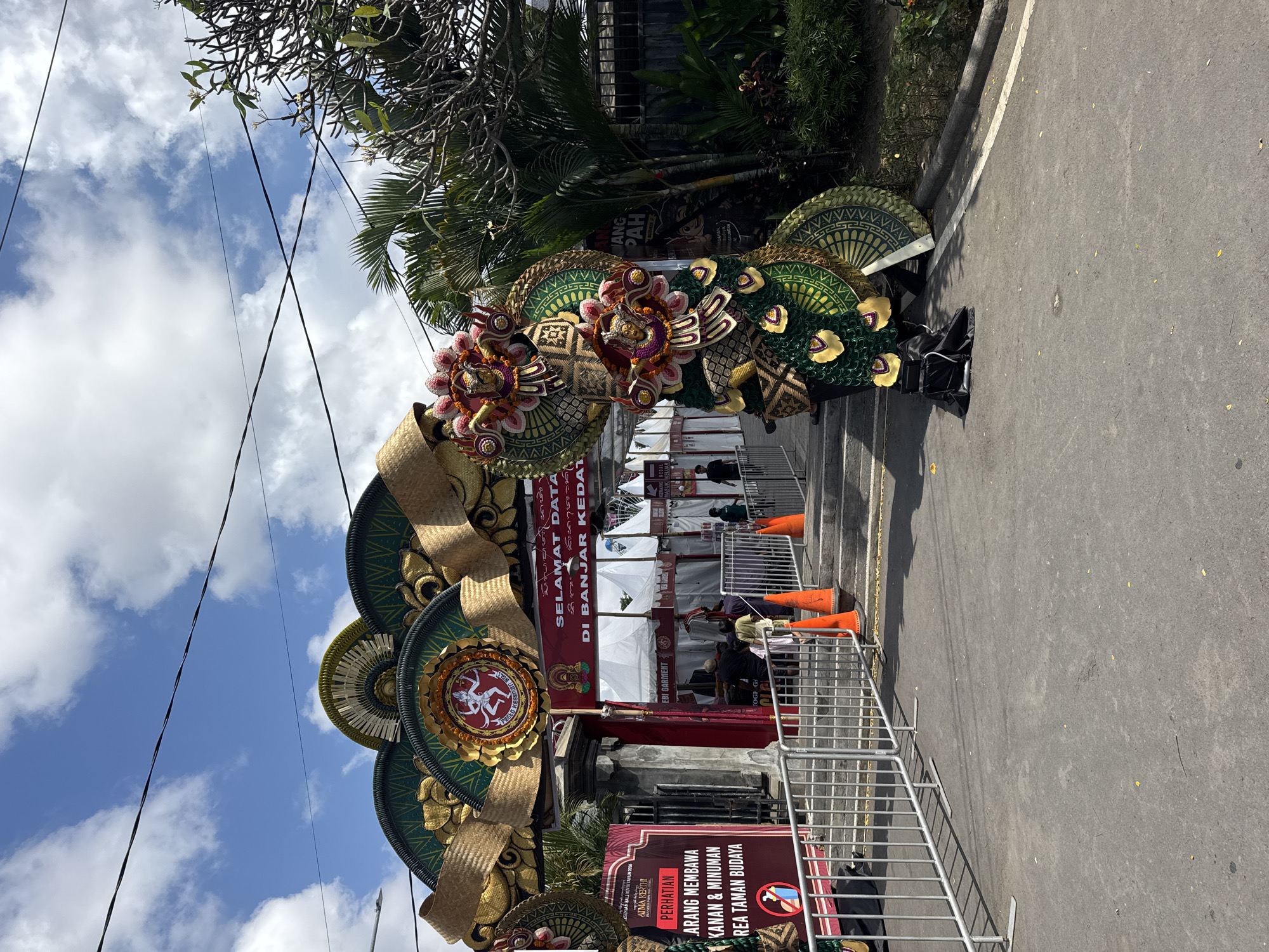 The image shows a street scene in Denpasar, Bali, Indonesia, likely near a cultural or festive event. The foreground features a paved pathway bordered by metal barricades on the left. To the right of the path, there are several orange traffic cones. Beyond the barricades and cones, a large, ornate archway serves as an entrance. The arch is decorated with intricate green and gold patterns, straw accents, and a central circular emblem featuring a dancing figure. Flanking the entrance are large, colorful, costumed figures that appear to be part of a parade or performance. These figures are adorned with vibrant green fabric, gold accents, and ornamental headdresses.
Under the archway, tents are set up, suggesting a market or exhibition area. A red banner with white text, reading "SELAMAT DATANG DI BANJAR KEDATON," indicates a welcome message and a specific location, "Banjar Kedaton," which is a traditional village administrative unit in Bali, often found within Denpasar. Some people are visible walking through the entrance, while others stand near the tents. Signs with Indonesian text are also present, one on the left bearing the word "PERHATIAN" (Attention) and further instructions. Palm trees and other lush vegetation are visible in the background, typical of Bali's tropical climate. The sky above is blue with scattered white clouds, and power lines crisscross overhead. The overall scene conveys a sense of an outdoor event or gathering, possibly a local festival or cultural exhibition in Denpasar.