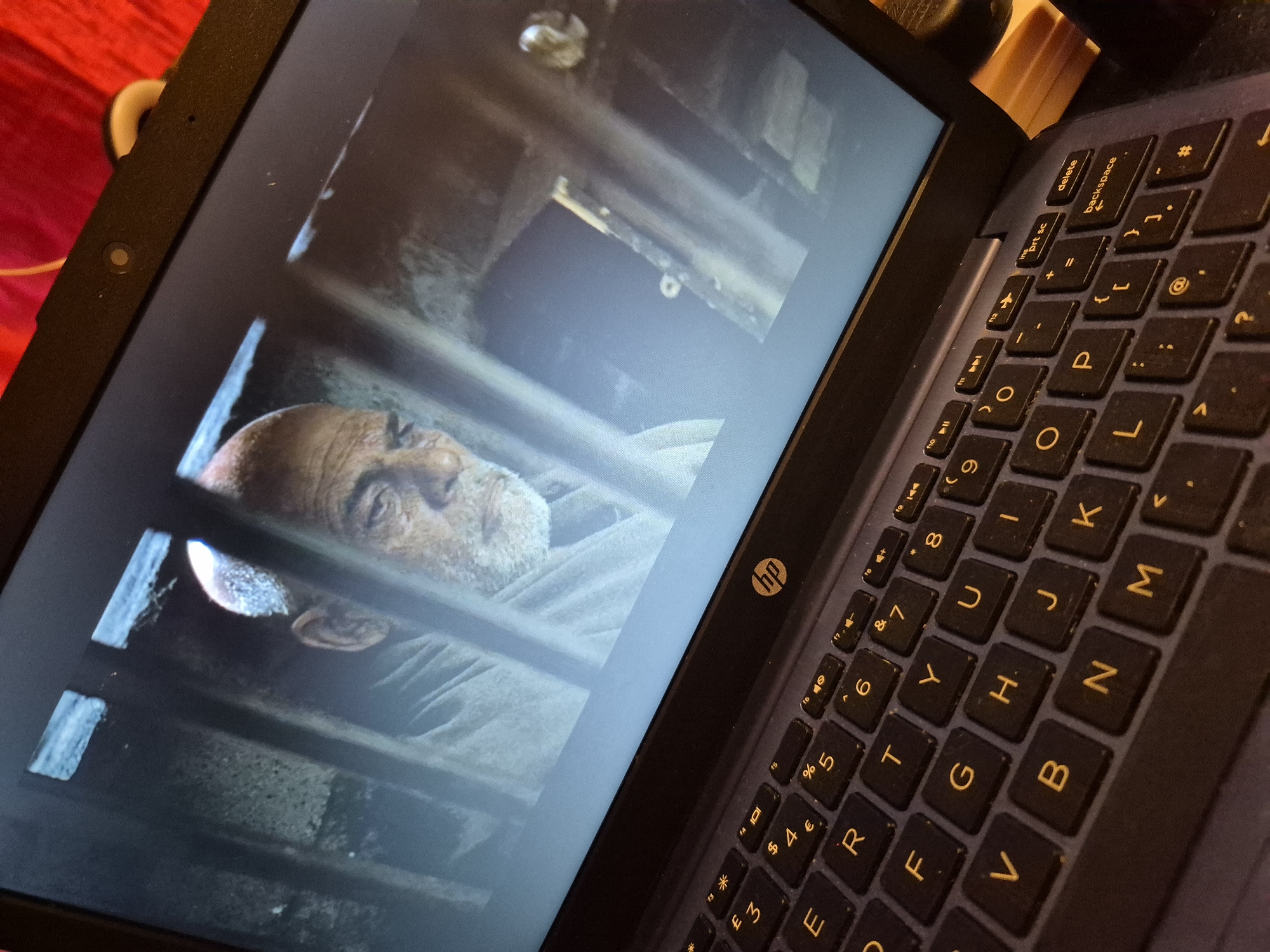 An elderly man with a beard and bald head appears on the screen of a laptop. The man is seen through bars, suggesting he is in a prison or similar confined space. The laptop keyboard is visible in the foreground. The image is taken from an angle that shows part of the laptop screen and the keyboard. The background of the laptop screen is dark and appears to be a still from a movie or TV show. The overall scene is a domestic setting, likely a home, with the laptop placed on a surface. There is no visible indication of the location within the city of Sandwell, United Kingdom.
