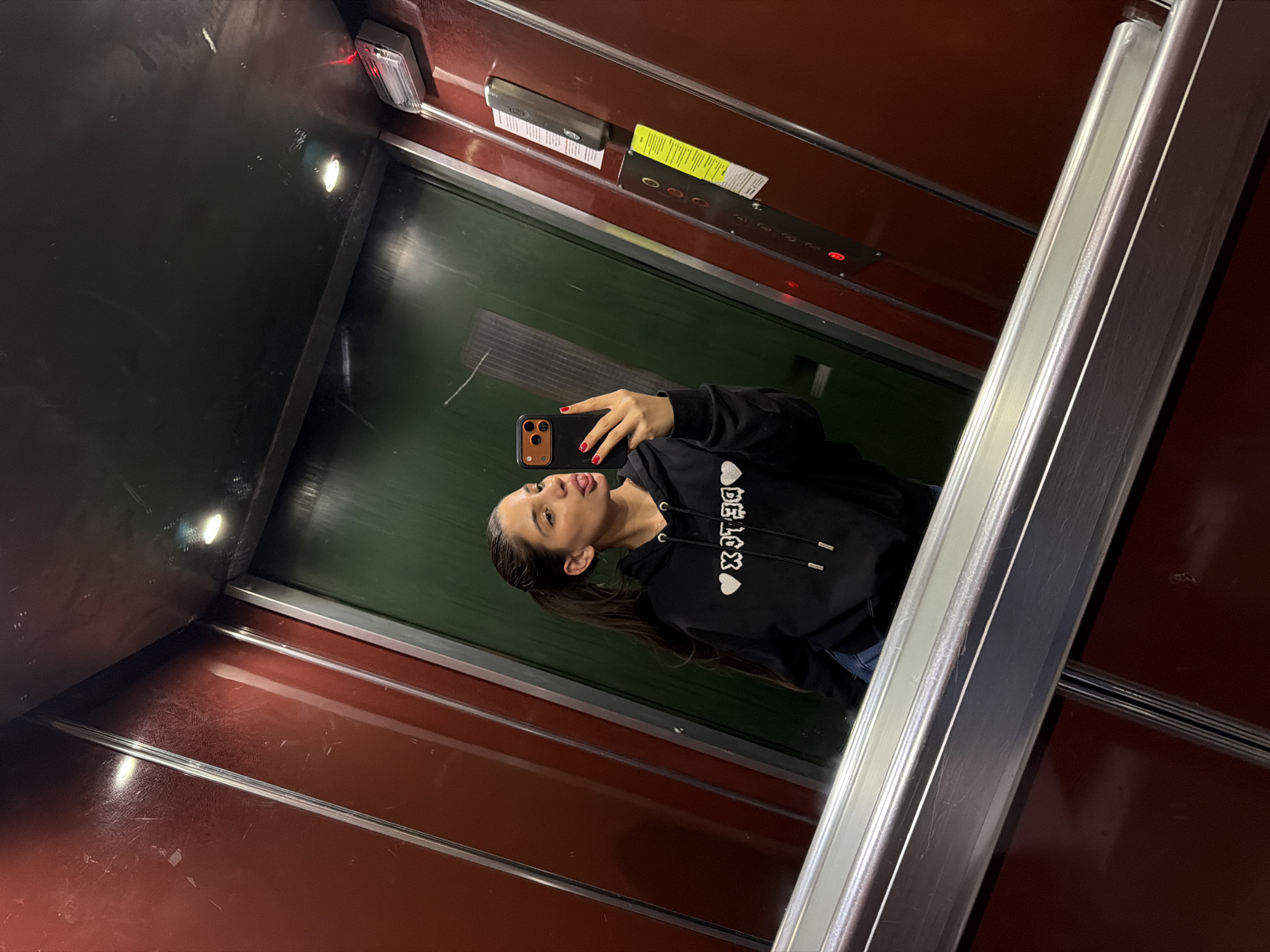 An adult female is captured in a mirror selfie inside an elevator. She wears a black hooded sweatshirt with reversed white text and heart symbols (appearing as "♡ DETOX ♡"), blue denim trousers, and red nail polish on her fingers. Her dark hair is pulled back into a ponytail. She holds a smartphone with an orange protective case in her right hand and protrudes her tongue. The elevator cab features dark reddish-brown and dark green wall panels, displaying multiple scratches and scuff marks, separated by vertical silver trim strips. A brushed metallic handrail, composed of two parallel bars, spans the lower portion of the foreground. On the right wall, a metallic control panel contains several pushbuttons, at least two of which are illuminated red, a speaker grille, a yellow instructional sticker, and a faded white information sheet. The dark ceiling contains a single recessed overhead light fixture emitting white light, and a separate emergency light unit with a red indicator is mounted on the right wall near the ceiling. The scene is situated within an elevator in Sofia, Bulgaria; however, no specific exterior landmarks or addresses are visible to pinpoint the exact location within the city.