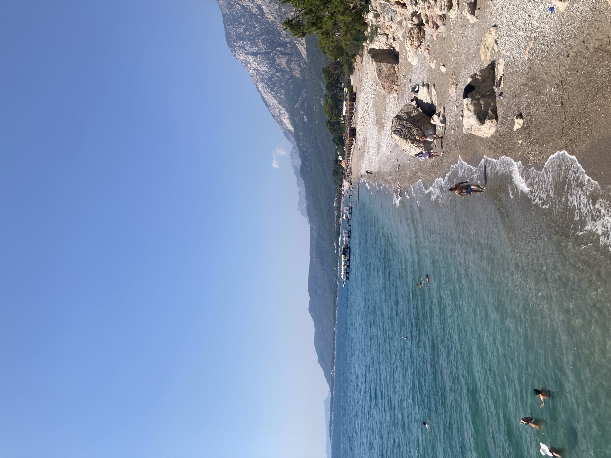 This is a daytime, eye-level, full shot of a pebble beach in Kemer, Turkey. The sky is clear and blue. The turquoise water of the sea is calm, with a few small waves breaking on the shore. Several people are swimming in the water. On the beach, there are sunbathers, a few people walking, and some straw umbrellas providing shade. In the background, there are mountains covered in trees, and a pier extends into the sea with people on it. The scene depicts a typical summer day at a Mediterranean beach resort.