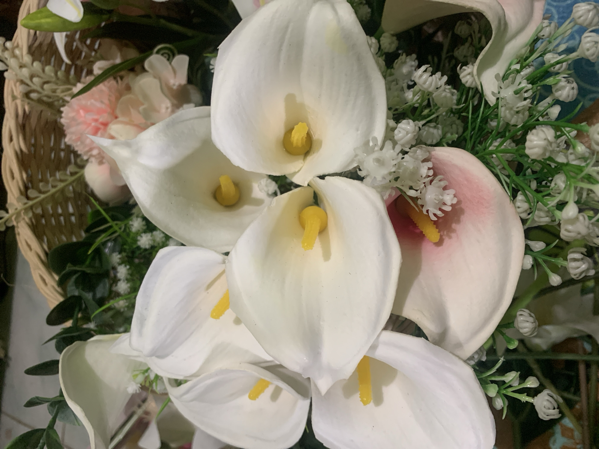 The image is a close-up of a bouquet of artificial calla lilies and other flowers arranged in a woven basket. The calla lilies are predominantly white with yellow stamen, and one appears to have pink tinges. Small white filler flowers and green foliage are interspersed throughout the arrangement. The background shows a hint of a tiled surface. There are no people or actions depicted. The location in Kuala Pembuang, Indonesia is not discernible from the image itself.