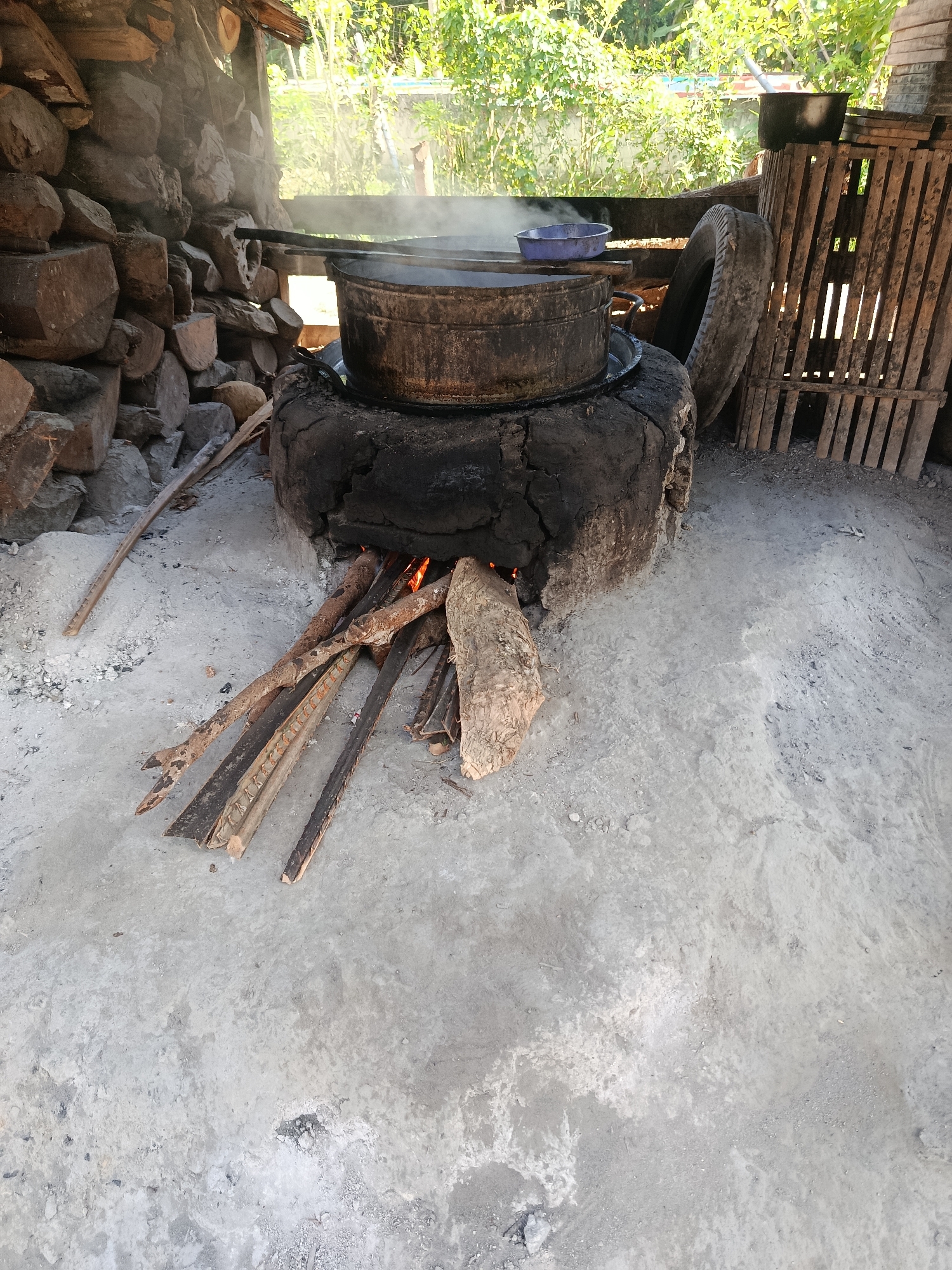 An outdoor scene depicts a makeshift cooking setup with a large pot steaming over a tire-based stove, likely located in Sarwadadi, Indonesia. A pile of firewood is placed in front of the stove, indicating active cooking. The background shows a rustic wooden structure and lush greenery, suggesting a rural or semi-rural environment. The ground is covered in dirt and ash. No people are visible in the image.