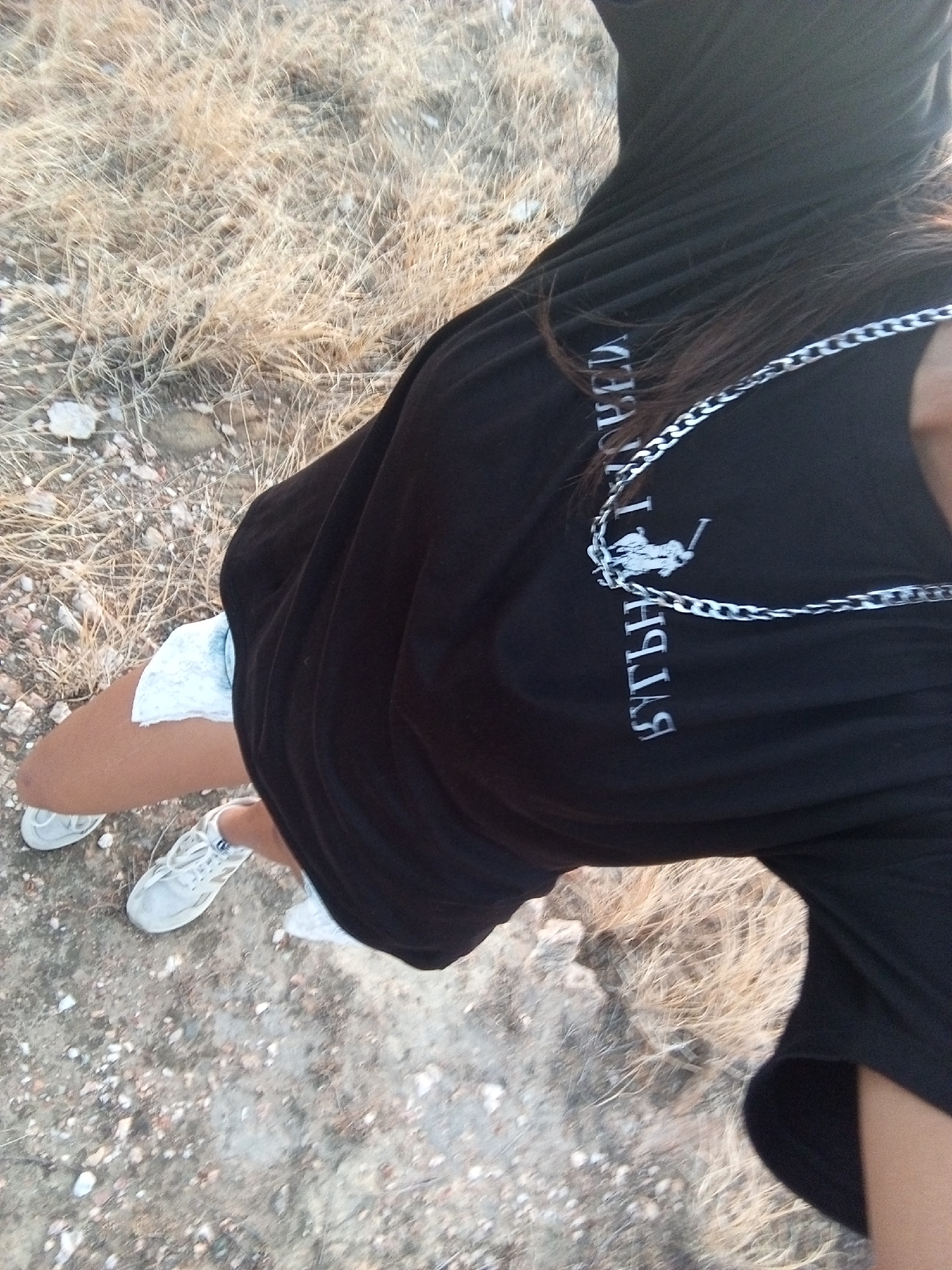 An overhead view captures a person standing on arid terrain in Juan Griego, Venezuela. The individual is wearing a black t-shirt featuring a white, inverted logo that includes a stylized polo player and accompanying text. A silver-colored chain necklace hangs around the person's neck. Light-colored (likely denim) shorts are visible on the lower body, paired with white athletic shoes. The ground consists of dry, light brown, sparse grass and scattered small rocks and dirt.