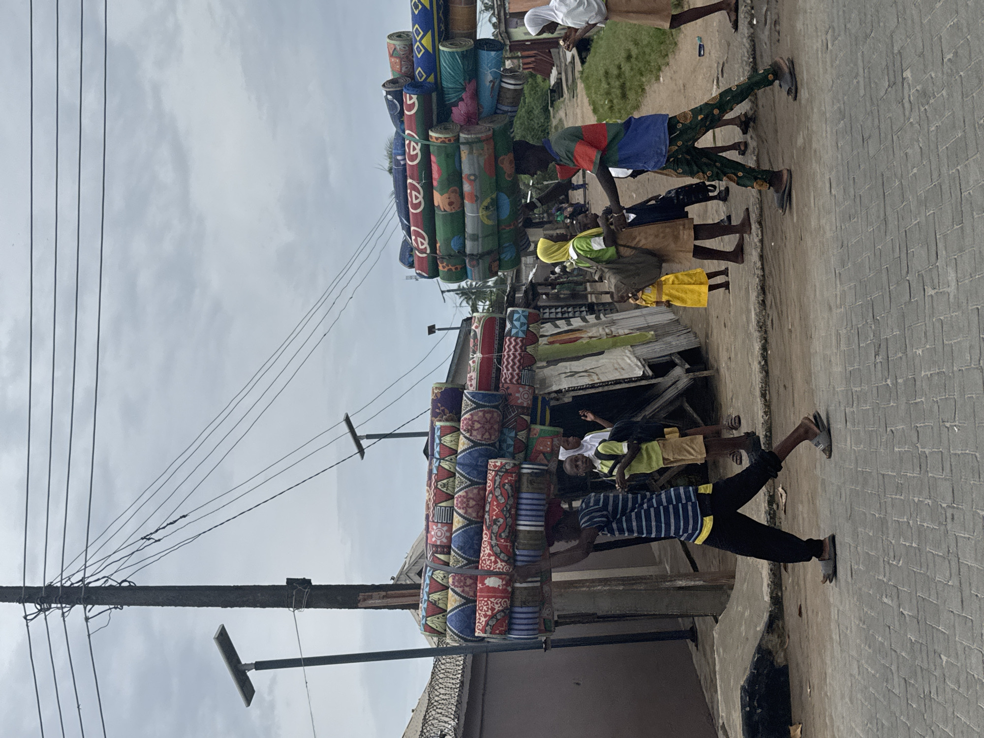 In Badagry, Nigeria, the image presents a street scene with people and stacks of colorful, rolled mats. A utility pole stretches into a cloudy sky, with several power lines connecting to it. In the foreground, a person in a striped shirt and dark pants is walking on a brick-paved road. Stacks of mats, featuring geometric and floral designs, are displayed near ramshackle buildings. Several people are gathered near the mats, including individuals in yellow and blue clothing. The setting suggests a local marketplace or commercial area.