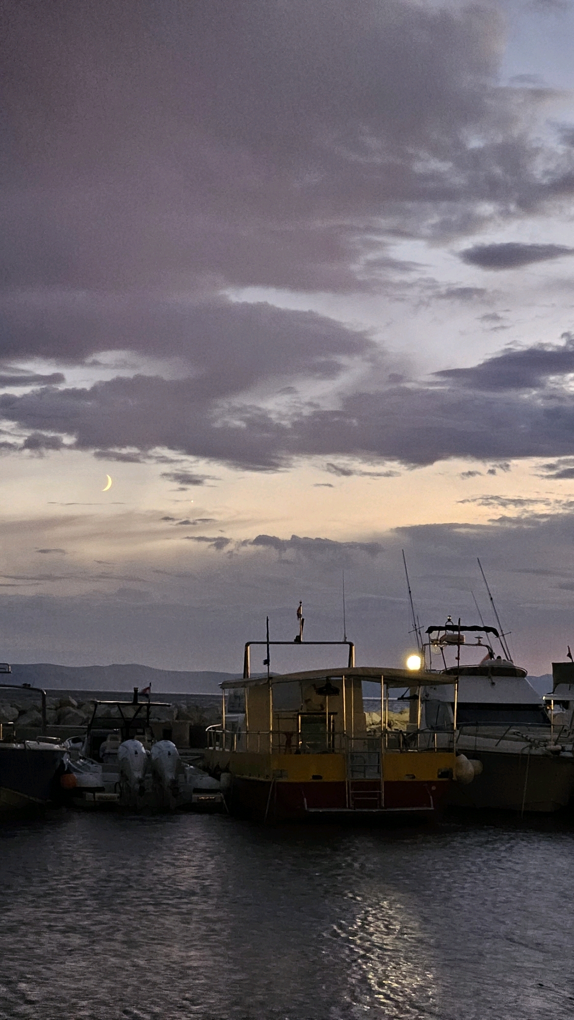A twilight scene shows boats docked in a harbor in Općina Tučepi, Croatia. The sky is filled with dramatic, striated clouds in shades of purple and gray, with a sliver of a crescent moon visible in the upper left quadrant. The water in the foreground is dark and rippled, reflecting the faint light of the setting sun and the artificial lights from the boats.
In the mid-ground, several boats are moored. The most prominent is a yellow and red boat with a covered cabin, illuminated by a bright light on its roof. To its left, another boat, possibly a dinghy, is visible. Further to the right, a white motorboat with fishing rods is docked. A rocky breakwater with a few sparse structures is visible on the left side of the image, separating the harbor from the open sea. In the far distance, a hazy, dark mountain range forms the horizon. The overall atmosphere is serene and dusky, with the last vestiges of daylight fading.