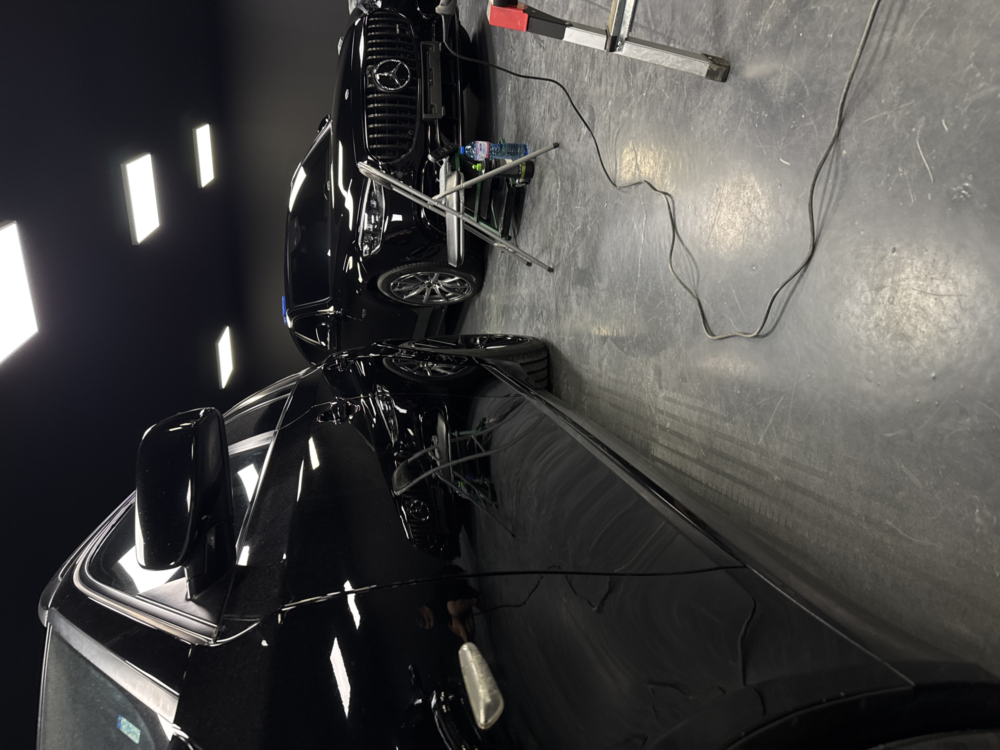 The image depicts the interior of a dimly lit garage or workshop, with a focus on two black cars. In the foreground, a portion of a black SUV is visible from a low-angle perspective, its glossy surface reflecting the overhead fluorescent lighting. Behind it, a black Mercedes-Benz sedan is parked, its grille and headlights illuminated. A foldable metal chair is positioned in front of the Mercedes, with a blue bottle of liquid and a spray bottle on a small cart beneath it. A power cord snakes across the concrete floor from the foreground towards the background, where a section of a silver ladder with red accents is also visible. A person, blurred due to motion, is crouched near the foreground car, seemingly working on it. The environment appears to be a professional detailing or auto repair space. The location is not specified in the image.