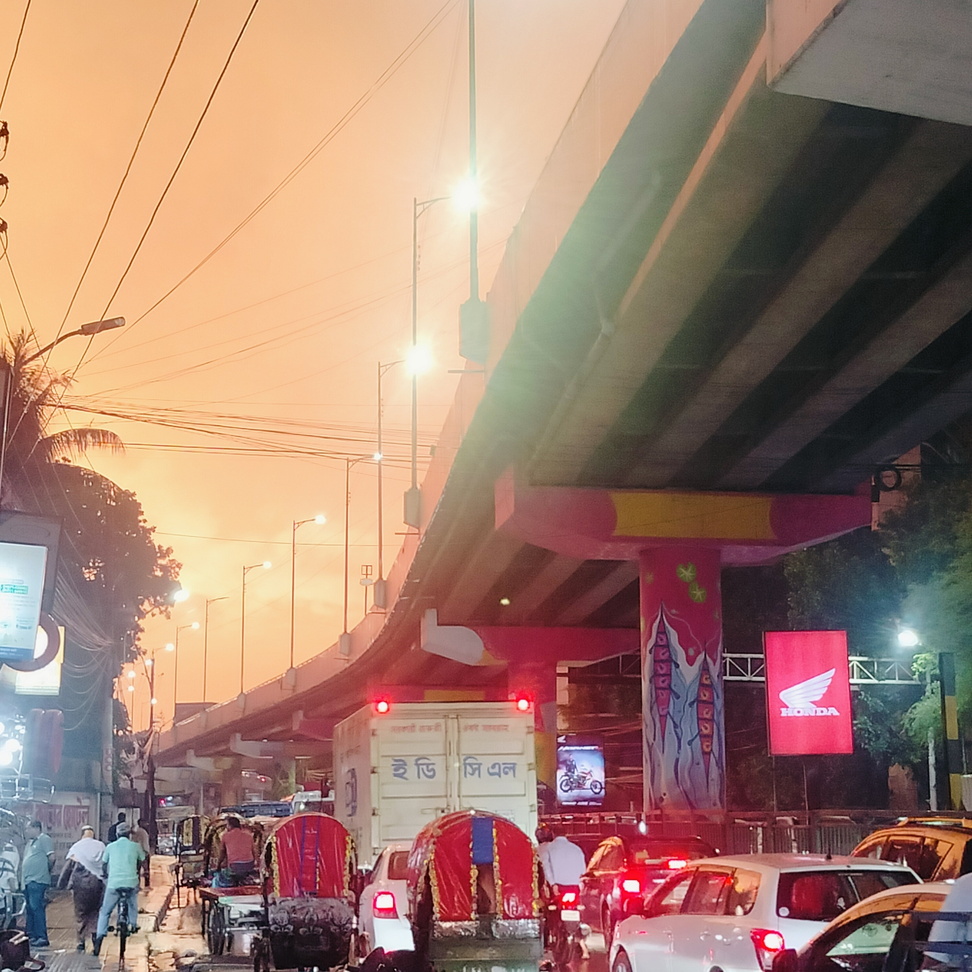 The image depicts a busy urban street scene in Dhaka, Bangladesh, during sunset or twilight. An elevated road or flyover dominates the upper portion of the frame. A Honda sign is visible, and the pillars supporting the flyover are painted with murals. The street below is congested with traffic, including cars, rickshaws, and motorcycles. Pedestrians are also present on the left side of the frame. The sky is orange, suggesting either sunrise or sunset. Streetlights illuminate the scene, and numerous overhead power lines are visible. A truck bearing the letters "ই ডি জি এল" (IDGL) is also visible.