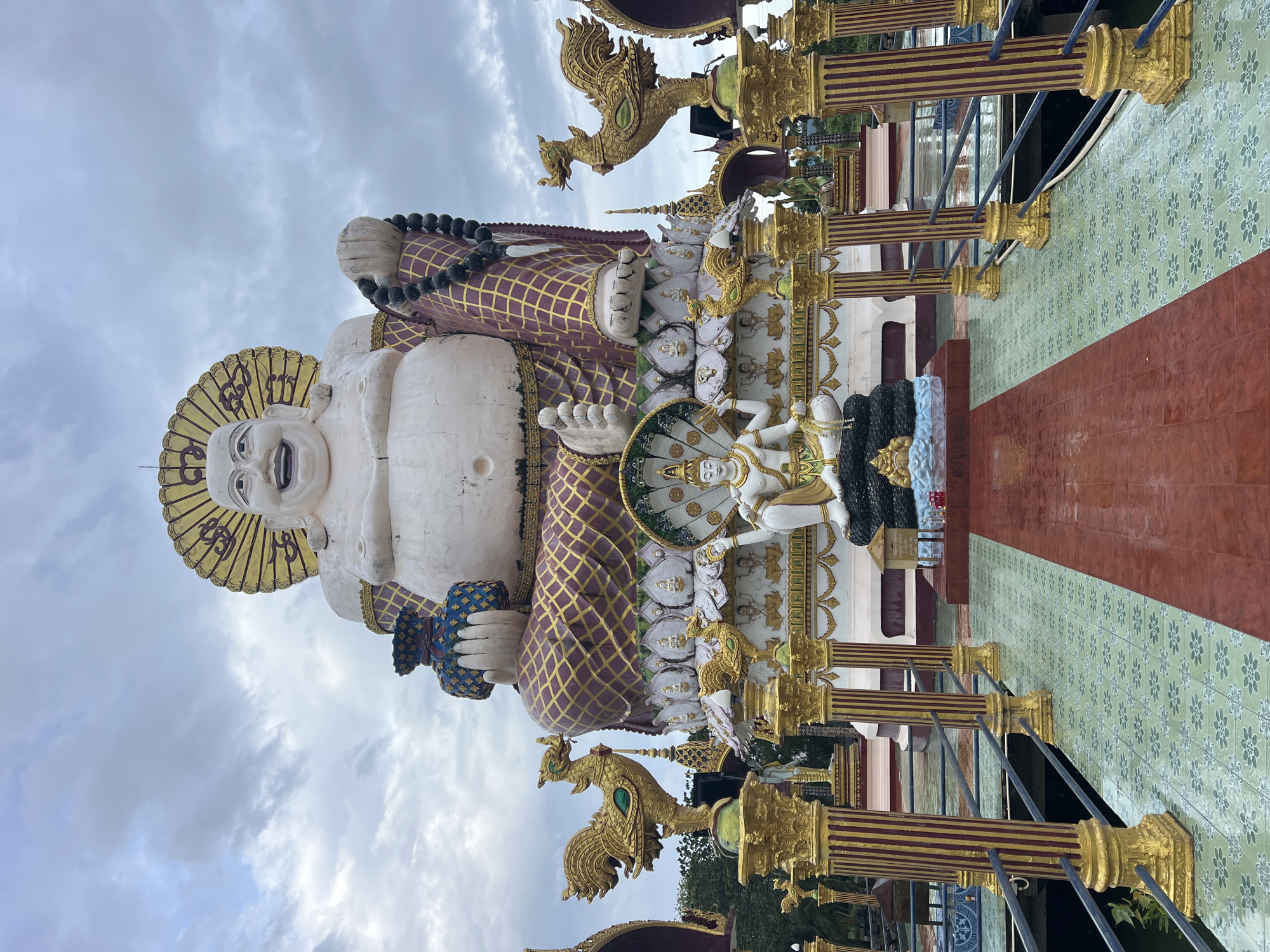 This image depicts a large, white statue of a smiling, corpulent figure, likely a representation of the Laughing Buddha, situated in the city of Baan Plai Laem, Thailand. The statue is adorned with a golden halo and wears a robe with a patterned design in purple and gold. It holds a blue vase in its left hand and a string of black beads in its right.
In front of the main statue, a smaller white statue depicts a seated figure with multiple arms, possibly a deity, resting on a coiled serpent. The area around the statues is decorated with ornate golden columns topped with bird sculptures. A pathway with patterned green and red tiles leads towards the statues, with metal railings on either side. The environment appears to be outdoors, with a cloudy sky overhead and greenery visible in the background. The overall scene suggests a religious or cultural site.