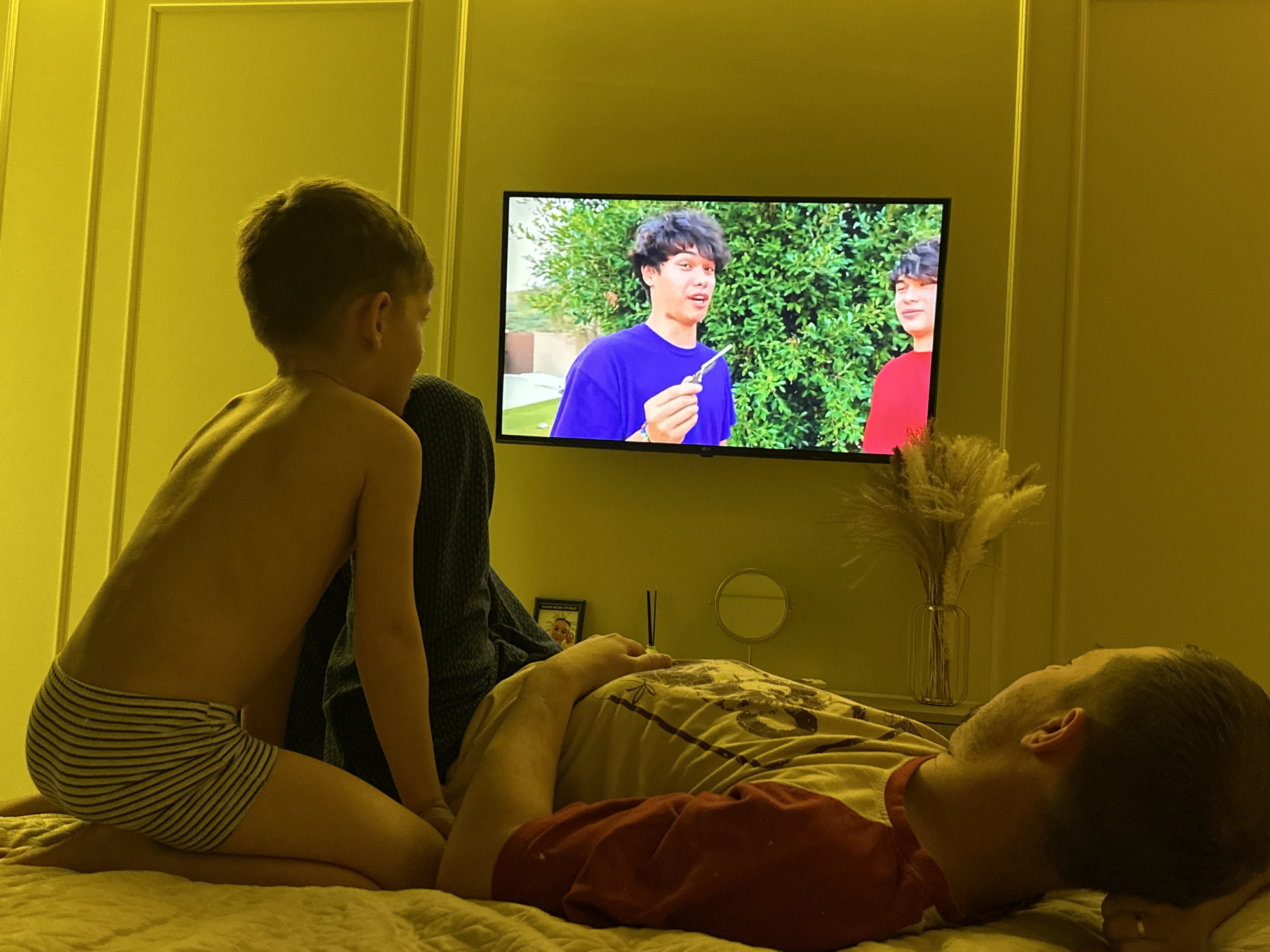 A young child in striped underwear is kneeling on a bed, looking at a television mounted on a wall. A man is lying on the bed. The television is playing a video of two young men standing in front of green foliage. There is a small framed picture, a small round mirror, and a vase of decorative grasses on a table near the television. The location may be a private residence in Chaikovsky, Russia.