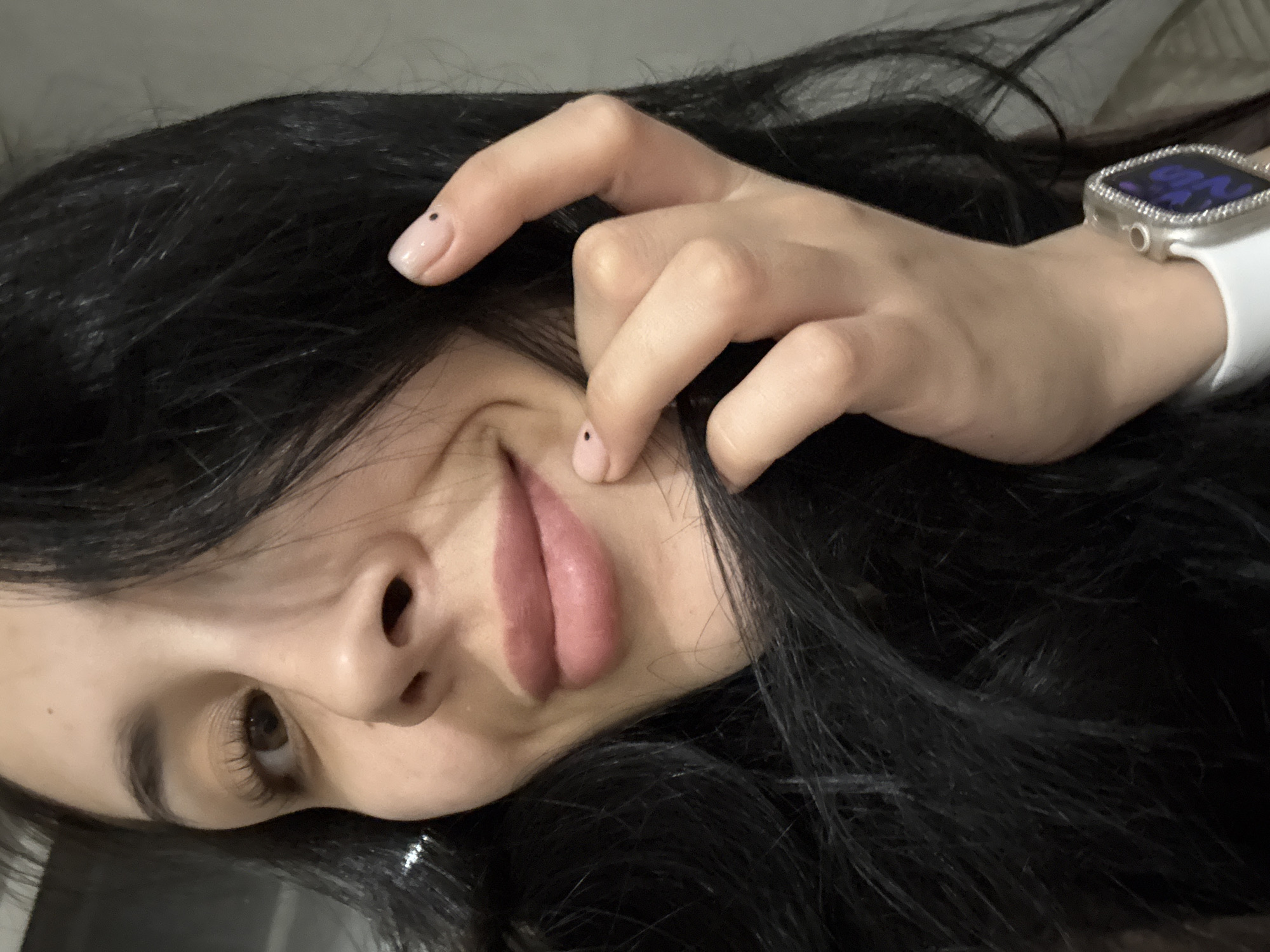 A close-up, face-down image captures a light-skinned person with dark, long hair and a focus on their face and hands. The person's face is angled slightly upwards, showing a single eye, nose, and lips. The hand closest to the camera has painted nails with a black dot on each. The person is wearing a watch on their wrist. No information indicates this photo was taken in Votkinsk, Russia.