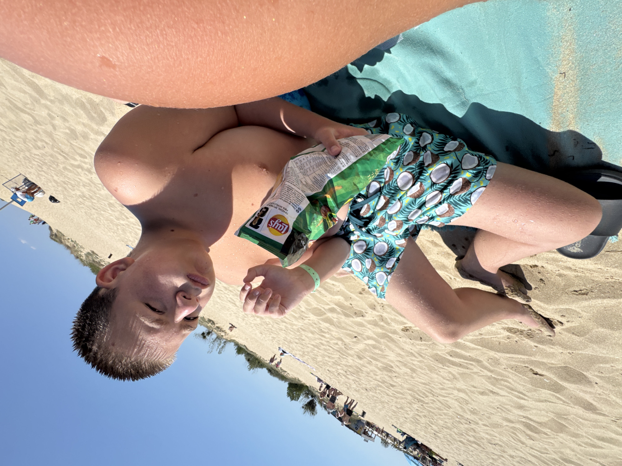 A low-angle, slightly tilted full shot shows a young boy sitting on a sandy beach in Nesebar, Bulgaria, holding a bag of Lay's potato chips. The boy is wearing teal swim trunks with a white coconut pattern. His skin is glistening with water droplets, suggesting he has recently been swimming. He is looking down at the chips and appears to be eating them. To the right of the boy, a portion of another person's arm is visible, with sun-kissed skin. The background reveals a crowded beach with beach umbrellas, people in the distance, and a sandy dune. The sky is a clear, bright blue. The overall impression is a relaxed summer day at the beach.