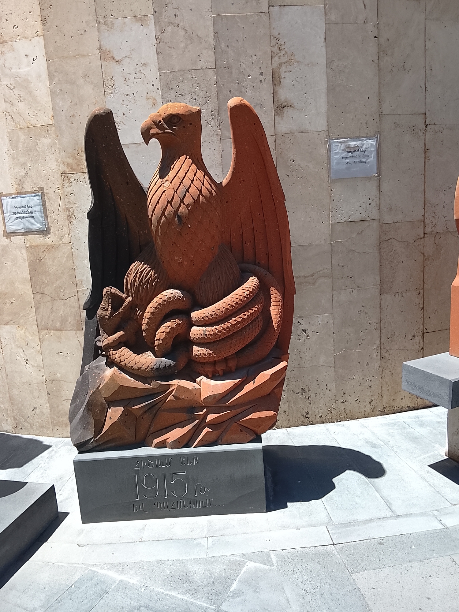 The image displays a reddish-brown, sculpted eagle grappling with a snake, placed outdoors against a light-colored tiled wall. The eagle, carved with detailed feathers and a strong beak, has its wings slightly spread, while the coiled snake is entangled at its base, suggesting a struggle or victory. The sculpture rests on a dark grey rectangular plinth.
The scene is brightly lit by natural sunlight, casting distinct shadows on the light-colored tiled ground, indicating daytime, likely midday or early afternoon. The overall setting appears to be a memorial or an outdoor exhibition area, possibly a museum garden, given the presence of similar bases for other, unseen sculptures.
Visible text on the main sculpture's base is in Armenian. It reads "ՀՐԱՆՏ ԷՐԱԶՄՈւՆԴ" (Hrant Erazmund, likely a name or dedication), followed by "1915 թ." (1915 year), and "ԵՎ ՊԱՀԱՆՋՈՒՄ..." (AND DEMANDING...). This explicitly references the Armenian Genocide, a significant event in Armenian history, which occurred in 1915, and implies a demand for recognition or justice.
Additionally, two laminated signs are affixed to the tiled wall, one clearly visible and the other partially obscured by the sculpture. Both signs bear the same Armenian text: "Խնդրում ենք արձանների վրա չբարձրանալ" (Please do not climb on the sculptures), indicating public access to the display and a cautionary instruction. The solemn theme of the sculpture, combined with the historical reference, suggests a place of remembrance and cultural significance.
