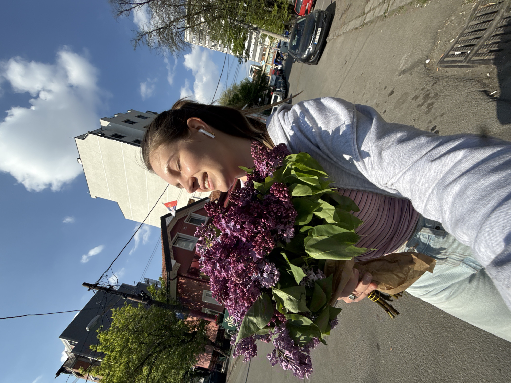 A vibrant, sunlit selfie-style photograph captures a young woman joyfully holding a large bouquet of purple lilacs on an urban street. The image is tilted, giving a dynamic perspective.
The woman, likely in her late teens or early twenties, is visible from the chest up. She wears a light grey hooded sweatshirt over a striped top. Her brown hair is tied back, and a white AirPod is in her right ear. Her eyes are closed or squinted in a broad smile, indicating happiness or amusement as she looks down towards the flowers. Her left hand, adorned with a ring, securely holds the base of the lilac bouquet, which is wrapped in brown paper. The flowers are a rich purple, accompanied by fresh green leaves.
The background reveals a bustling street scene under a bright blue sky dotted with scattered white clouds. To the left, a tall, modern white apartment building with multiple windows dominates the skyline. To the right, an older, red-brick house with white window frames and a red-blue-white striped flag (likely Serbian) is visible. A utility pole with wires stands between the buildings. The street features asphalt pavement and a concrete sidewalk, where a metal drain grate and scattered cigarette butts are visible. Several cars are parked along the street, including a black car with a distinct Serbian license plate "BG 031 YA", which confirms the location as Belgrade, Serbia. Partially visible on the red-brick house is a green sign with white Cyrillic text, reading "ПАО...". Trees with green foliage and some bare branches line the street.
The photograph conveys a sense of lightheartedness and the simple pleasure of a sunny day with beautiful flowers in an urban setting, specifically within the Vozdovac Urban Municipality in Serbia.