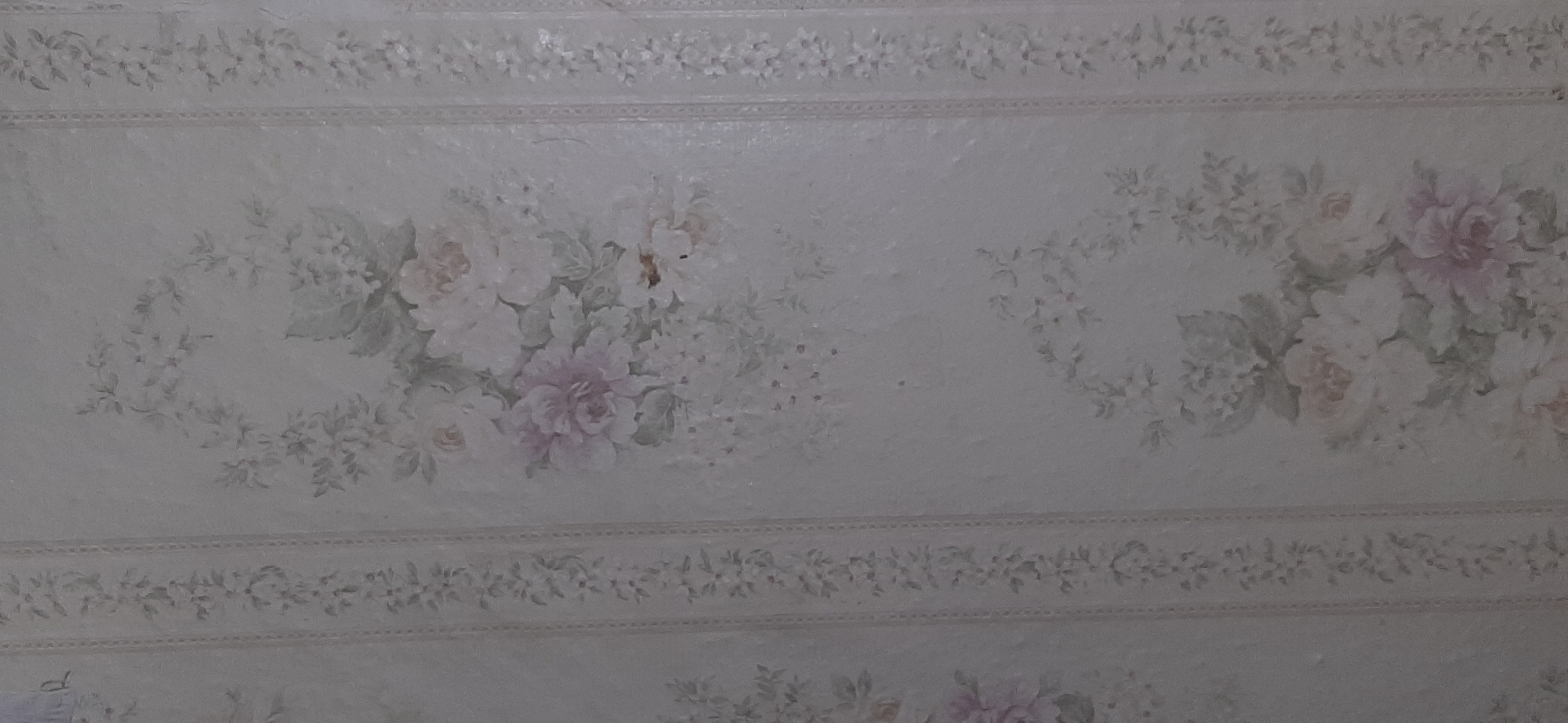 The image shows a close-up of floral wallpaper. The wallpaper has a light, off-white background with a subtle texture. There are vertical border stripes with delicate floral patterns. The main floral motifs appear to be large, soft-petaled flowers in shades of white, pink, and lavender, interspersed with smaller blossoms and green foliage. The style is classic and somewhat vintage. The image does not provide any information about a specific location in Yessentukskaya, Russia, or any people, actions, or interactions. It is solely a detailed view of a wall covering.