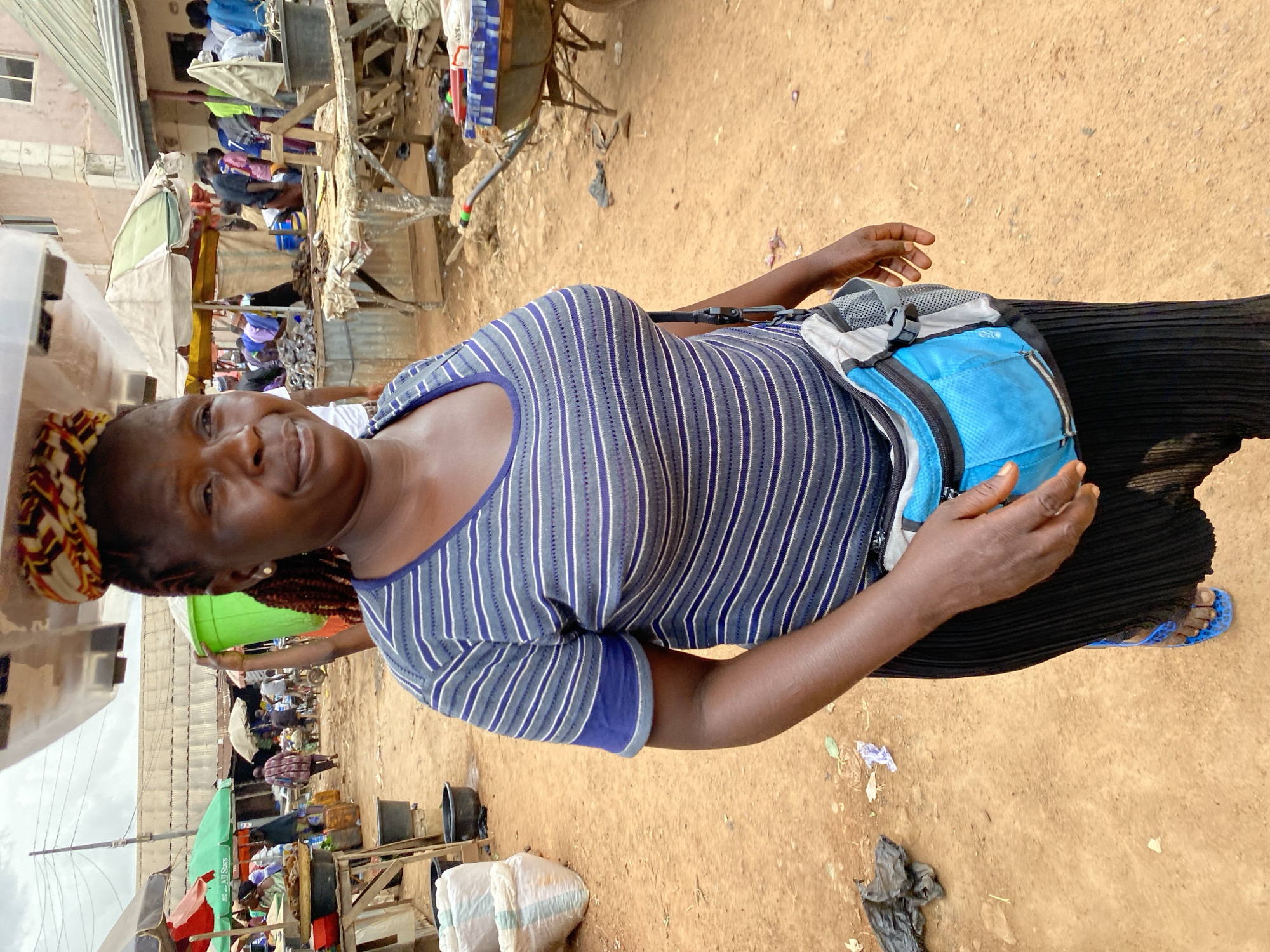 A Black woman stands in a bustling outdoor market in Bukuru, Nigeria, carrying a large, white, rectangular object balanced on her head. She is wearing a striped blue and white t-shirt, a black skirt, and blue flip-flops. A blue and gray fanny pack is worn around her waist. In the background, various market stalls with awnings and goods are visible, along with other people shopping and working. The ground is covered in reddish-brown dirt.