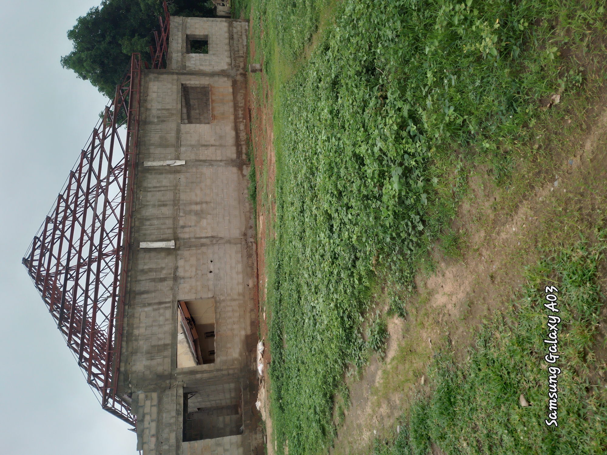 The image depicts an unfinished building with a red metal roof structure in Doka-Tofa, Nigeria. The building is constructed from gray concrete blocks, and its roof is exposed, revealing the metal framework. Openings for windows are visible, some with boarded-up sections. A second, smaller structure with a brown roof is partially visible inside one of the openings. Surrounding the building is an overgrown area with green vegetation and a dirt path. Two white sacks are placed on the ground near the building. In the background, a large green tree stands tall. The sky is overcast and gray. The lower left corner of the image displays the text "Samsung Galaxy A03".