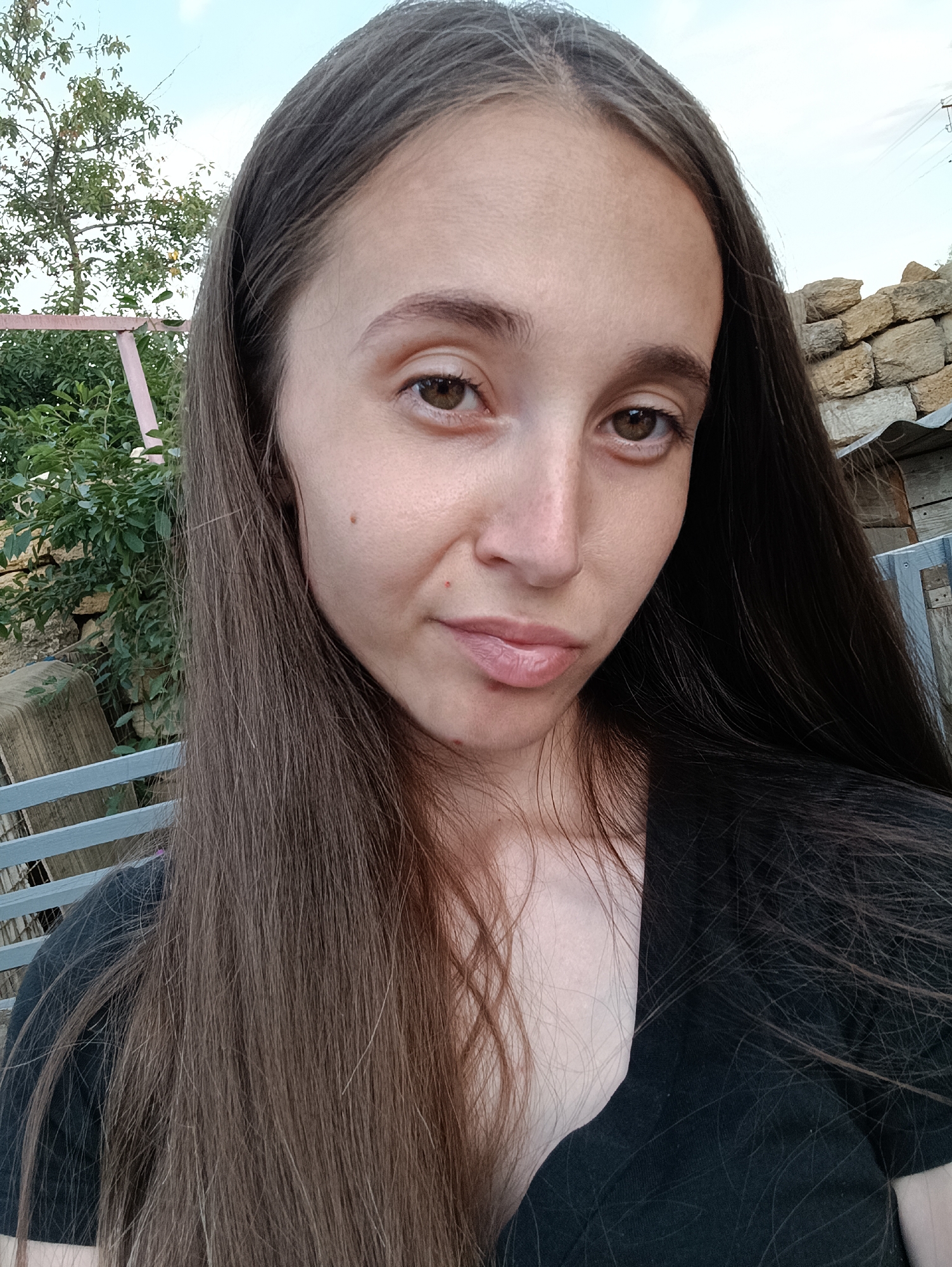 A close-up, waist-up, outdoor selfie taken from a slightly low angle features a young adult with long, straight, dark brown hair. She has fair skin, visible moles on her cheek, and brown eyes. Her lips are slightly parted, and she is wearing a black shirt. The background reveals a rustic setting with a stone wall, wooden structures, greenery, and a glimpse of a pale blue sky. The environment suggests a rural or semi-rural location in Vypasne, Ukraine.