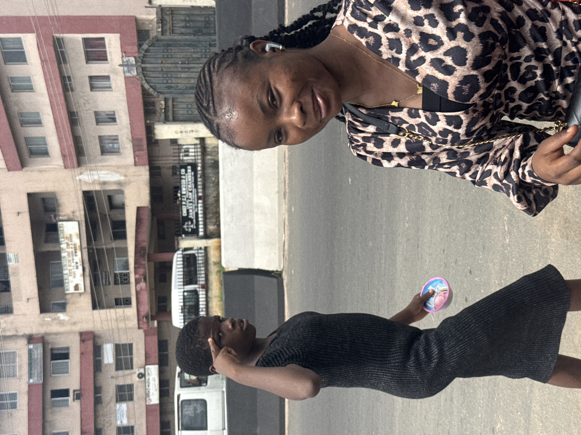 In Owerri, Nigeria, two young women are captured in a medium shot on a city street. The woman on the left, with short, closely cropped hair, is wearing a dark, ribbed, sleeveless dress. She is shown in profile, looking to her left with her right index finger raised to her forehead, as if in thought or saluting. In her left hand, she holds a small, round, colorful object. The woman on the right is looking towards the camera, with her head tilted slightly. She has braided hair and is wearing a leopard-print top with a low neckline and a black camisole underneath. A gold chain with a star pendant is visible around her neck. Both women appear to be walking on a paved road, with a grey concrete barrier behind them. In the background, a multi-story building with multiple windows and a large, ornate metal gate are visible. A white van is parked in front of the building, and there are signs on the building, one of which reads "NIGERIA UNION OF TEACHERS". The overall scene suggests a candid moment captured in an urban setting.
