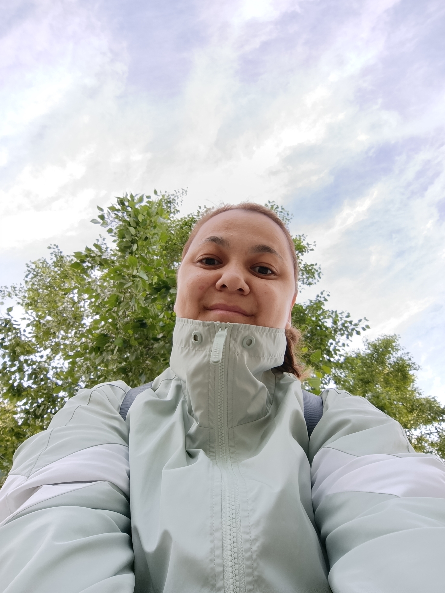 An eye-level selfie shows an adult individual looking directly into the camera, positioned at the upper center of the frame. The person has light brown hair, pulled back, and is wearing a light mint green or pale teal zip-up jacket with a high collar, which features white or light grey panels on the shoulders and upper arms. A grey strap, likely from a backpack, is visible over their right shoulder. The person has a neutral or slightly pleasant expression.
The setting is outdoors, likely in a park or natural area in Chaikovsky, Russia. Behind the individual, dense green foliage from trees occupies the lower half of the background, extending to both sides of their head. Above the trees and the person's head, a bright sky with scattered white and light grey clouds is visible. The lighting suggests it is daytime, with soft, diffused light, indicating partly cloudy weather. The photograph is taken from a low angle, looking upwards towards the subject and the sky. No discernible text is visible within the image.